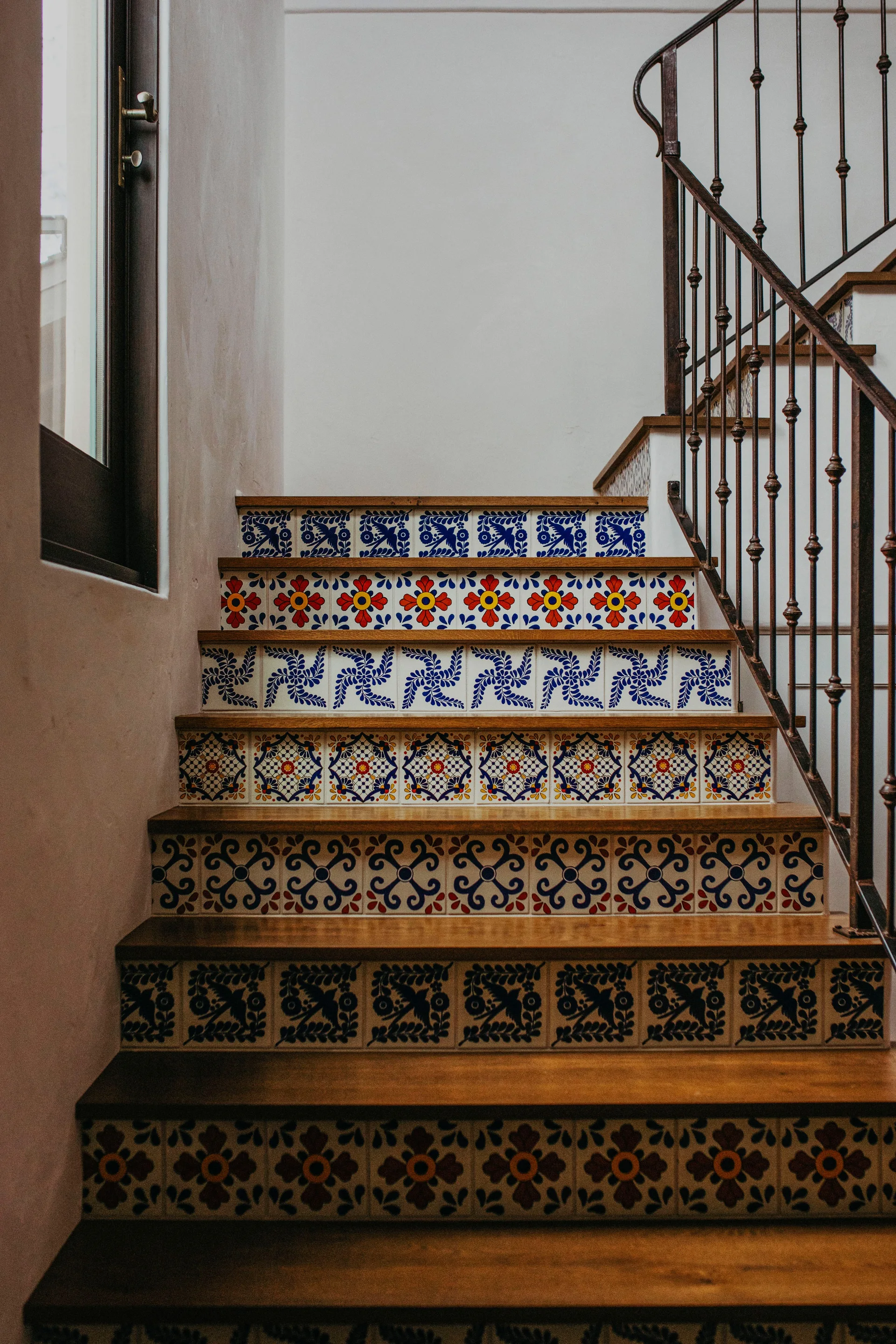 An indoor staircase features dark wood treads, a wrought iron railing, and colorful patterned Mediterranean tiles on each riser.