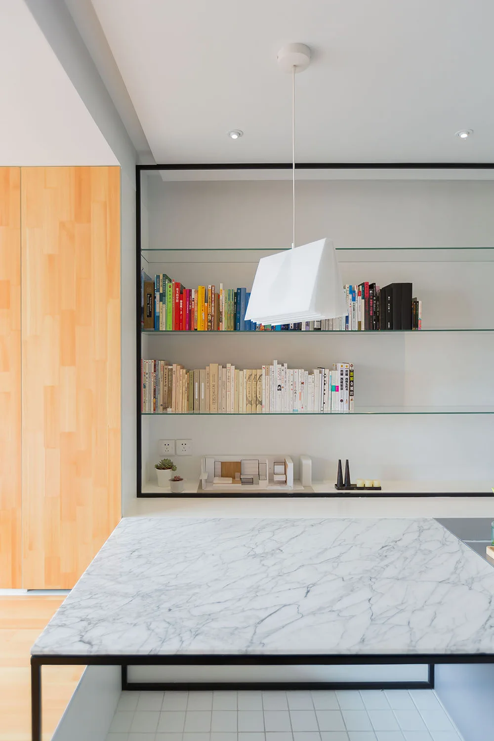 A minimalist interior features a white veined marble island below a white geometric pendant light and glass wall shelves lined with colorful books.