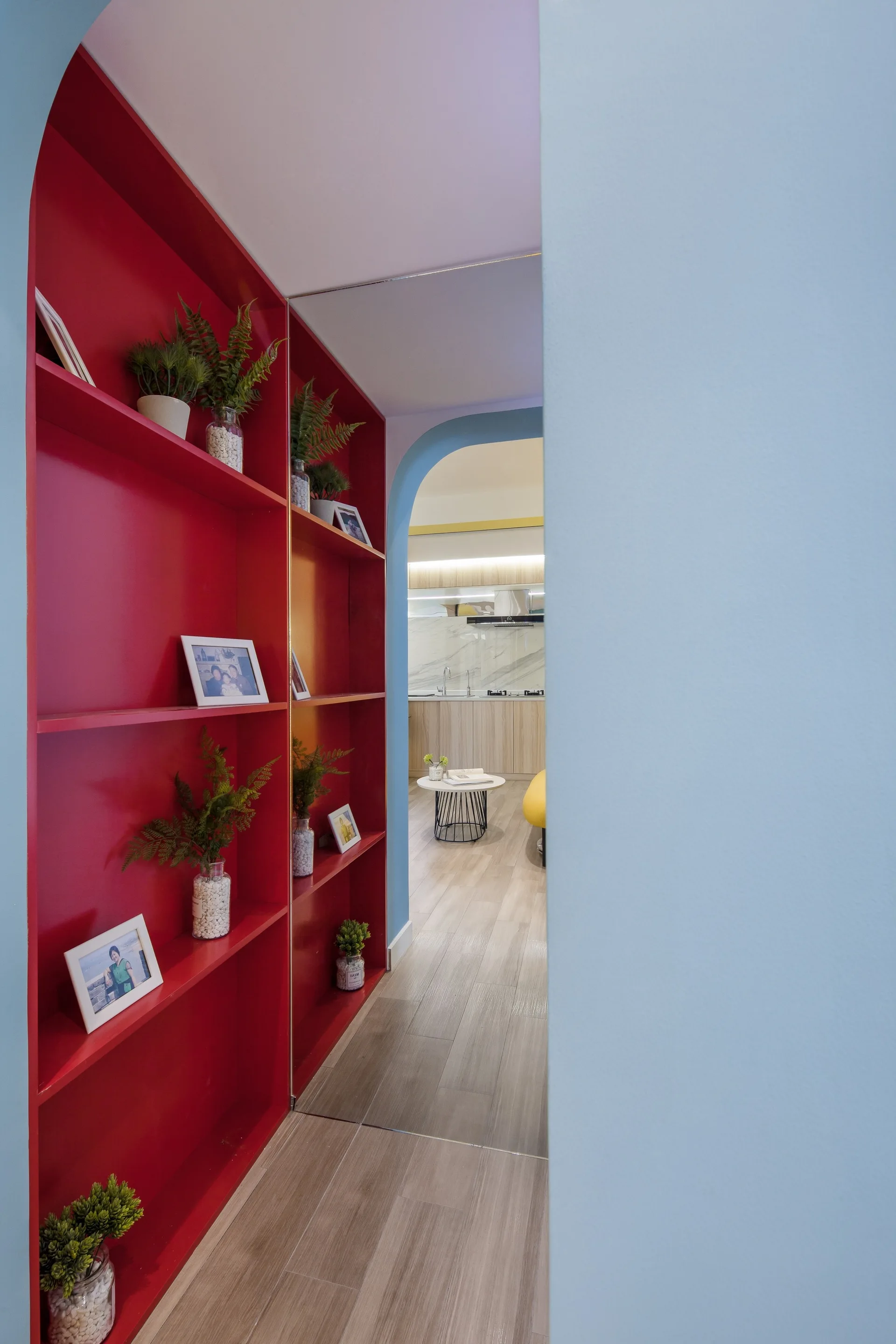 A light blue hallway featuring a bold red built-in shelving unit decorated with plants and frames, leading through an archway.