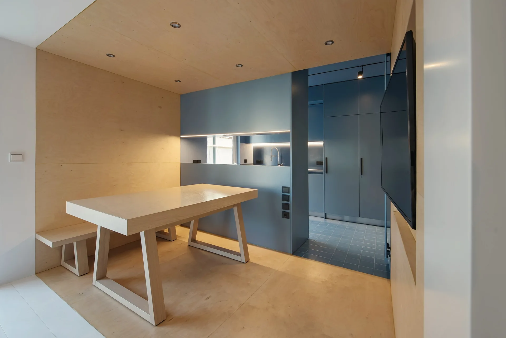 A minimalist dining nook with a light wood table, bench, and paneled ceiling sits next to a matte blue kitchen partition wall.