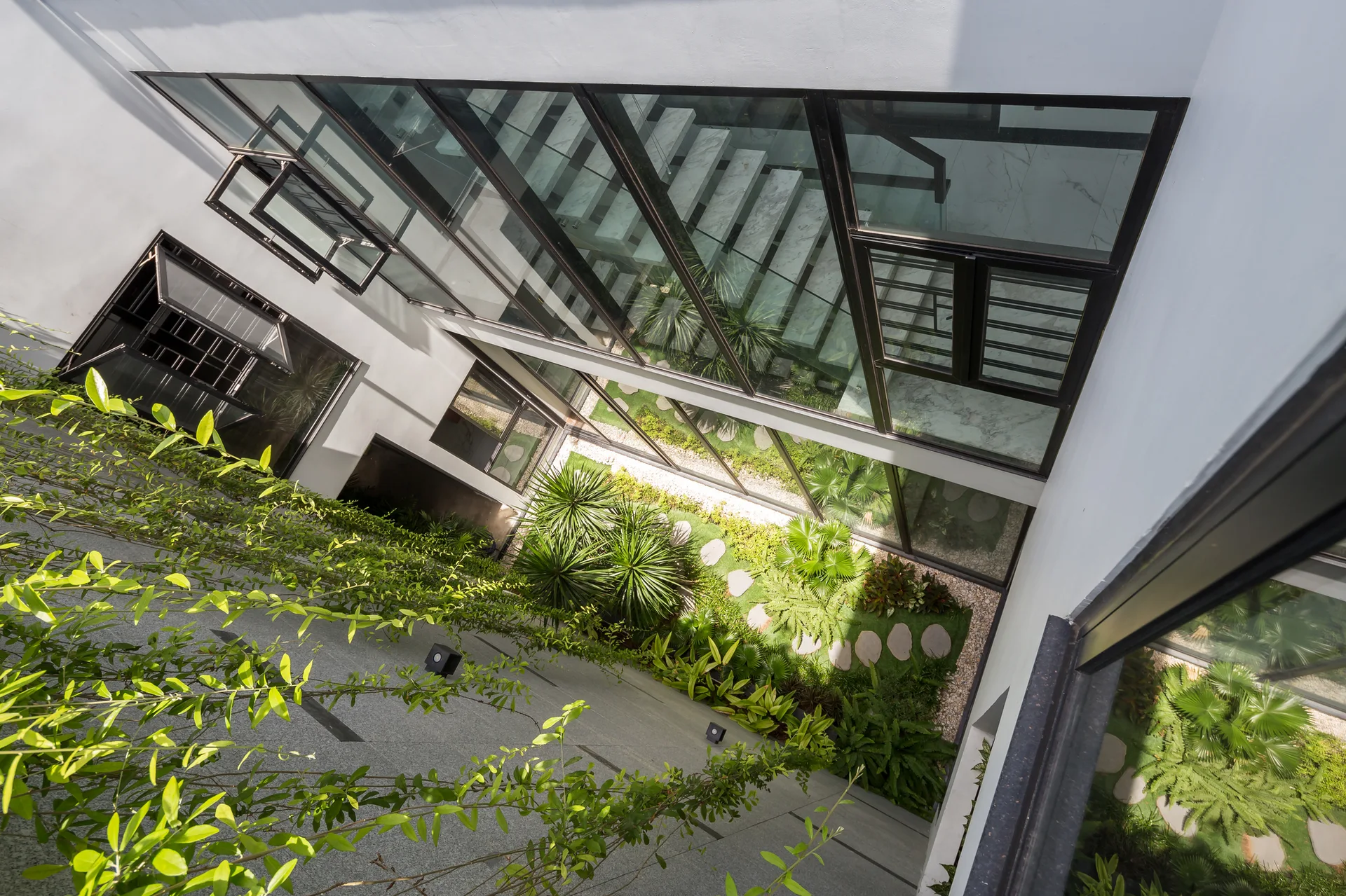 A high-angle view of a modern interior courtyard garden featuring green plants, stepping stones, and a glass-enclosed staircase.
