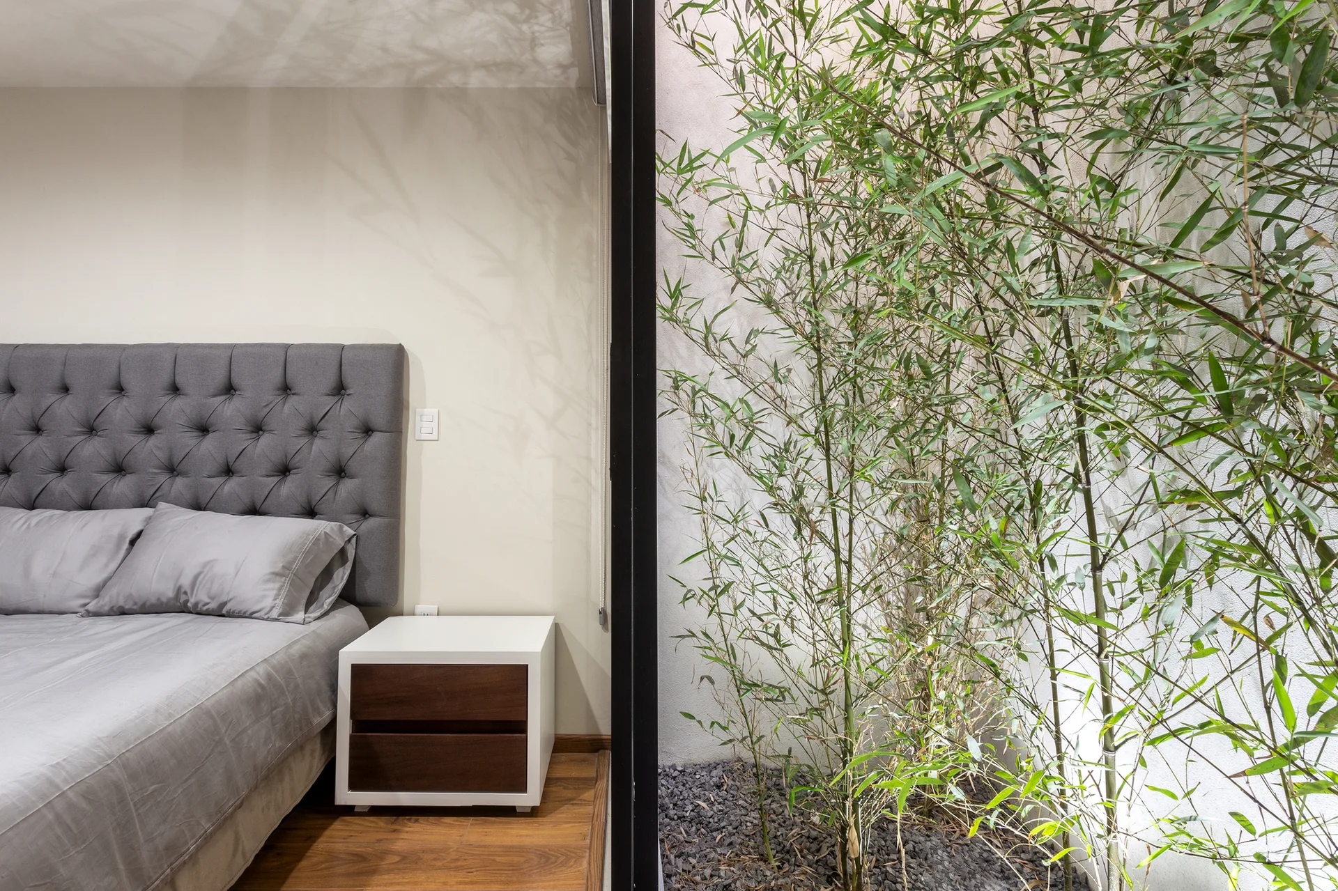 A modern bedroom features a grey tufted headboard and two-tone nightstand next to a glass wall showing an indoor lightwell with green bamboo.