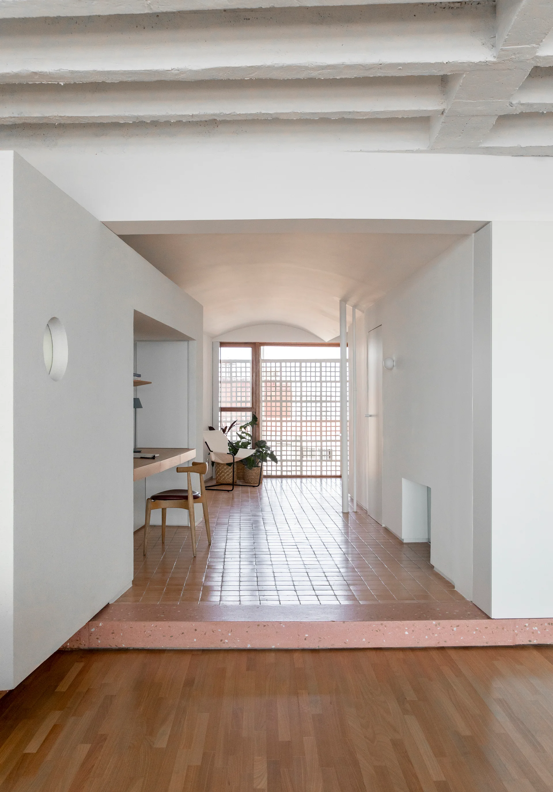 A minimalist corridor features a pink terrazzo step transition to terracotta tiled floors, a built-in wooden desk, and a vaulted ceiling.