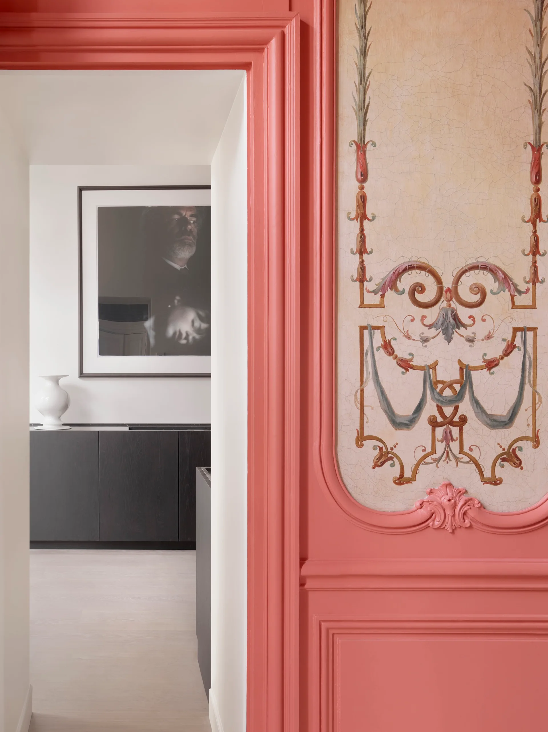 A bright coral door frame with ornate moldings stands next to a painted wall mural, framing a view of a minimalist hallway with black cabinetry.