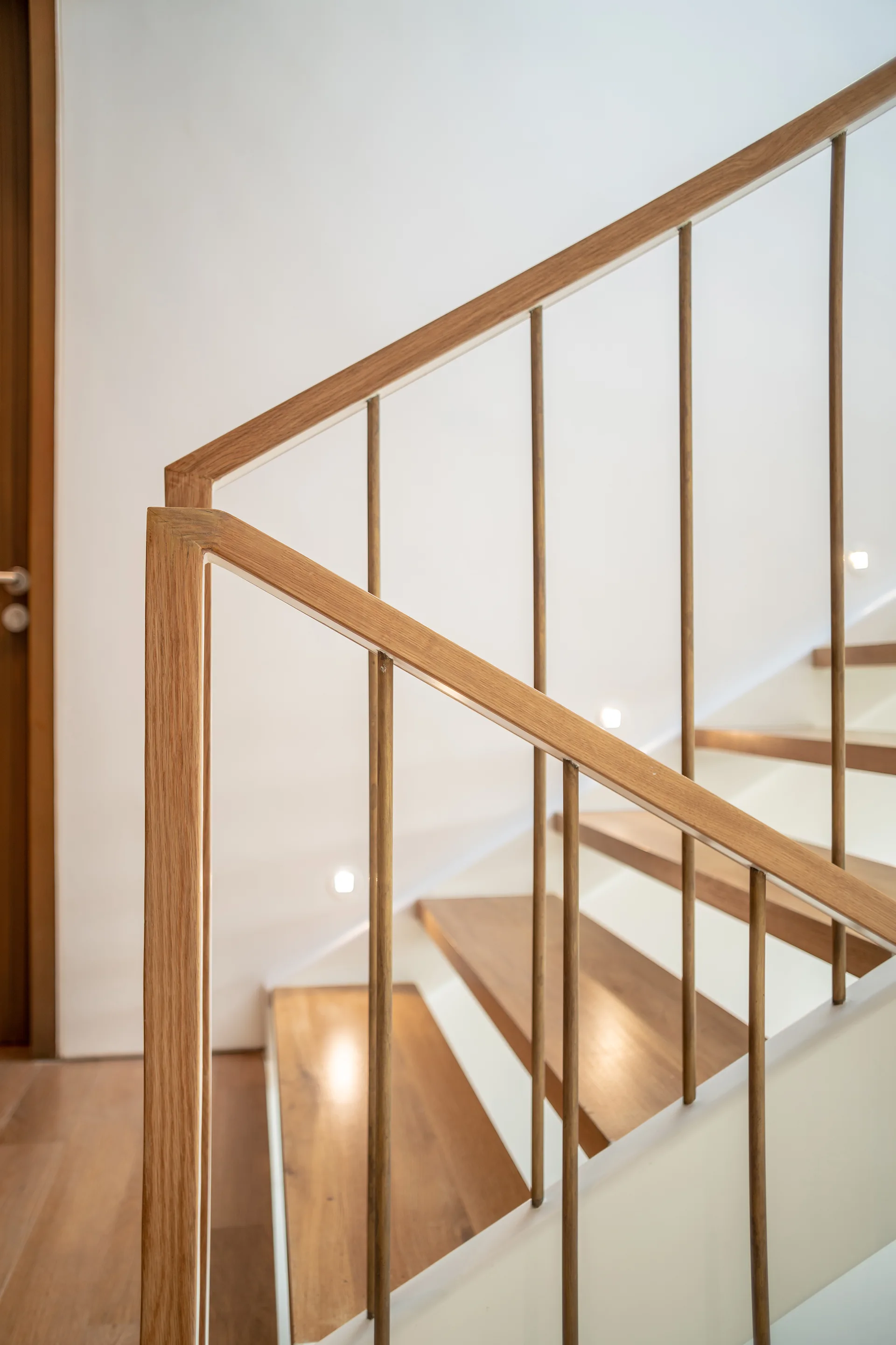 A close-up of a minimalist staircase featuring light oak wood treads, matching wood handrails, and thin metal spindles against a plain white wall.