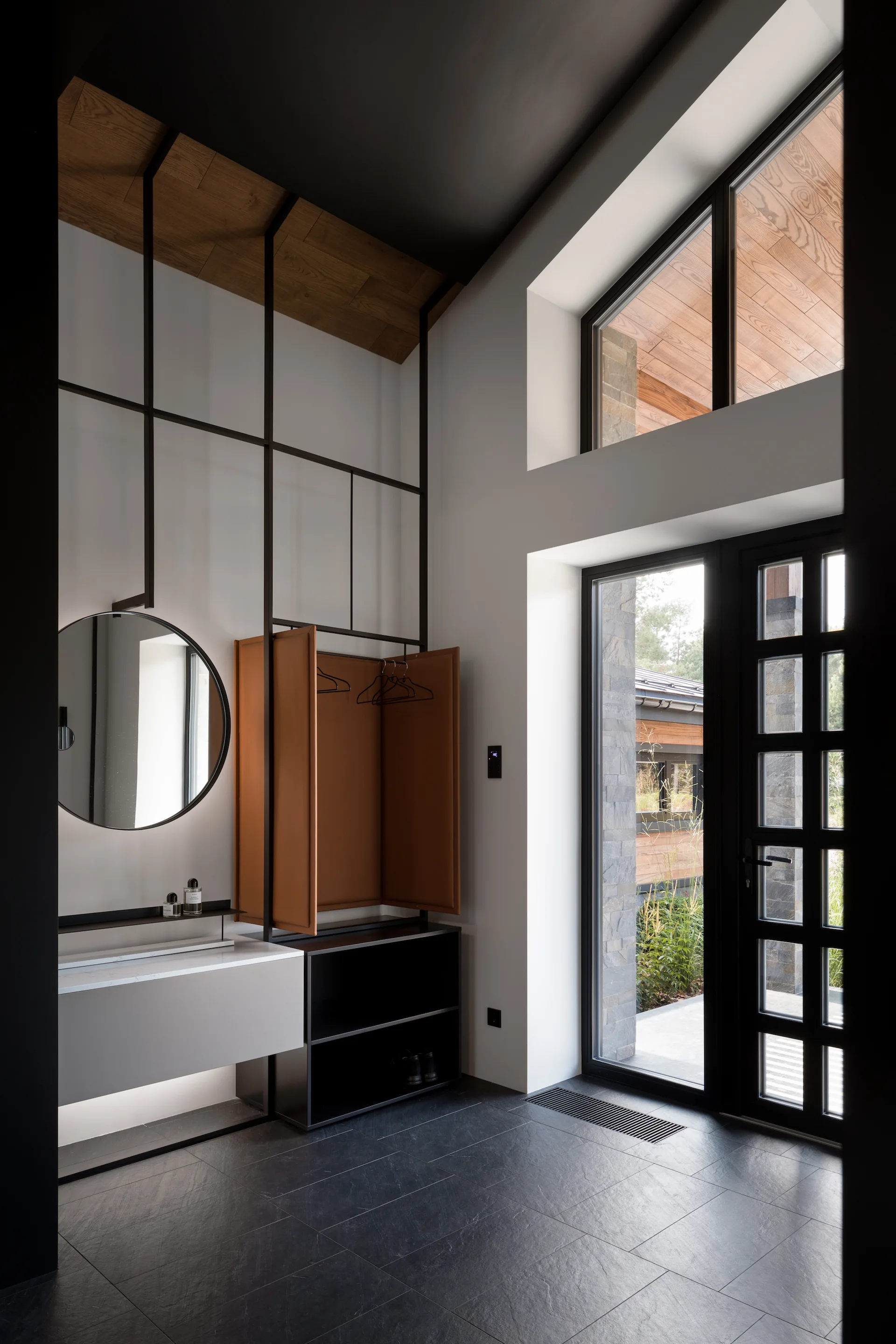 A minimalist residential entryway features a black-framed coat rack, a wall-mounted sink vanity with a round mirror, and a black grid glass door.