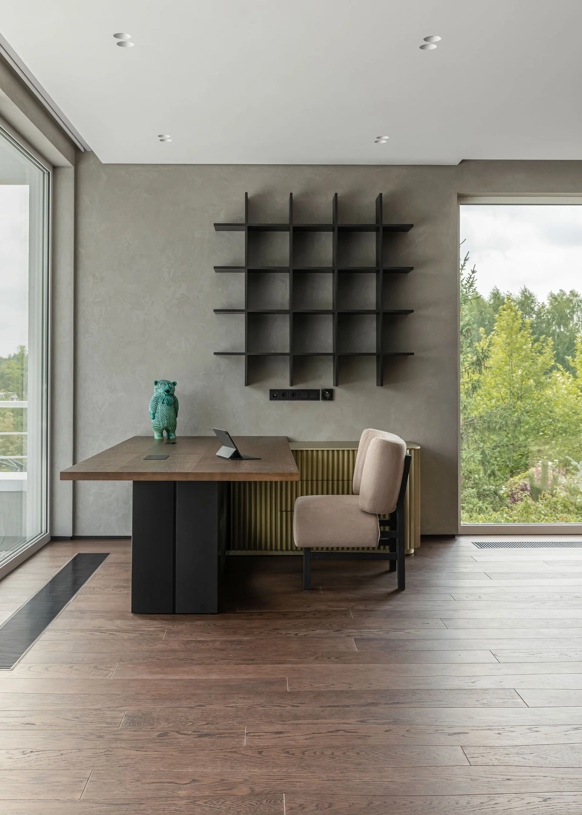 A minimalist home office displays a wooden desk, a beige upholstered chair, and a black grid shelf mounted on a textured grey wall.