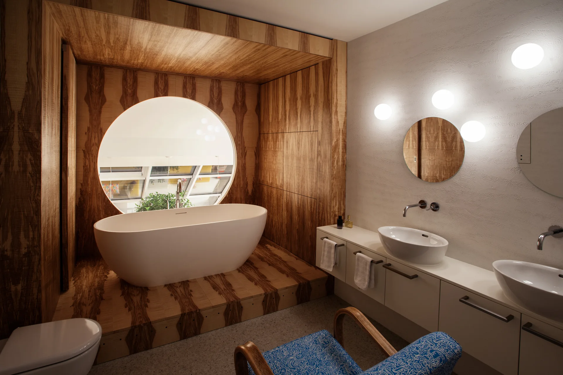 A white freestanding bathtub sits on a raised wood-paneled platform inside a matching wood alcove with a large circular window.