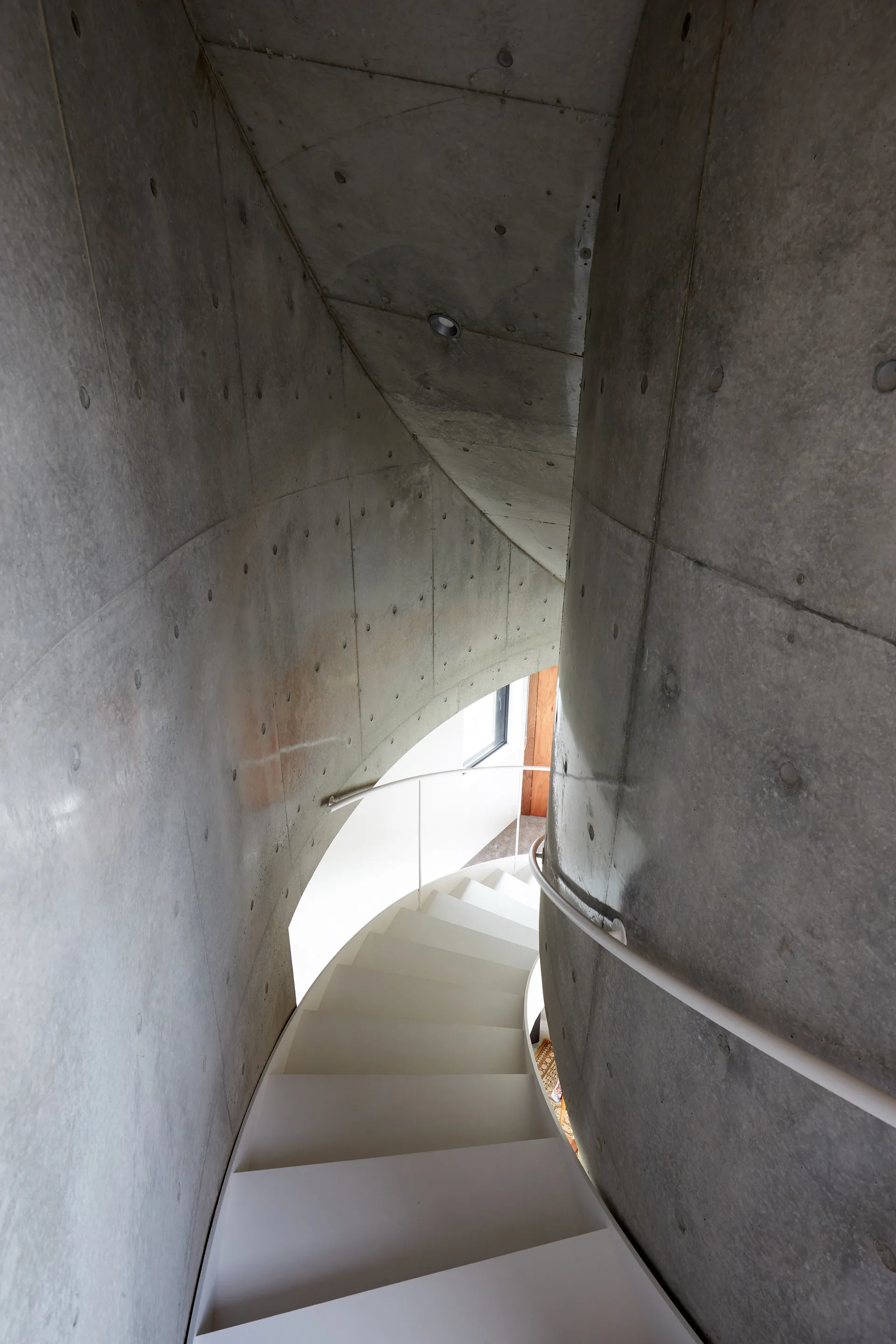 A winding white spiral staircase curves downward between two massive, smooth grey concrete walls with visible structural markings.