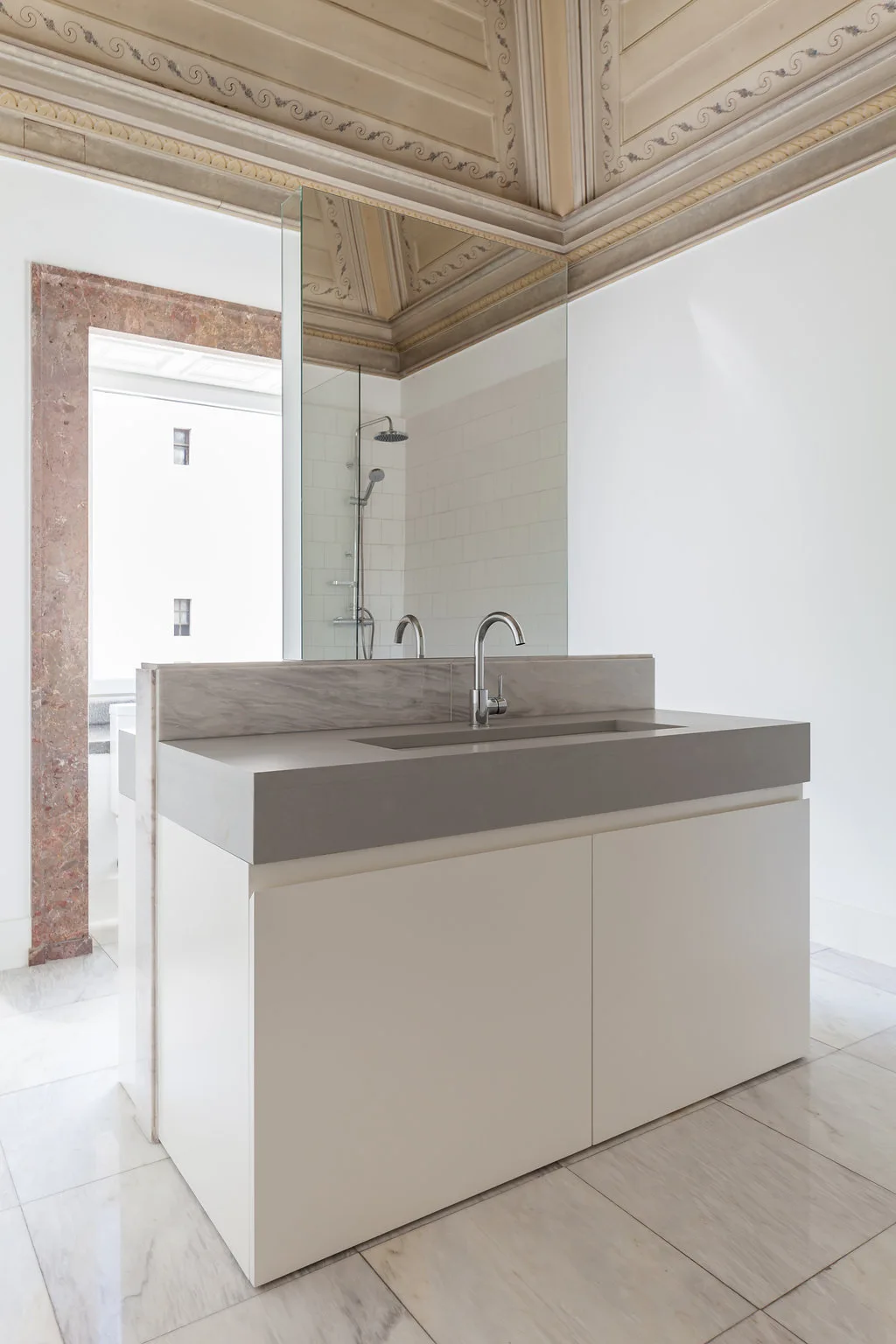 A minimalist white vanity with a gray stone countertop sits in a bathroom featuring an ornate painted ceiling, marble floor, and large mirror.