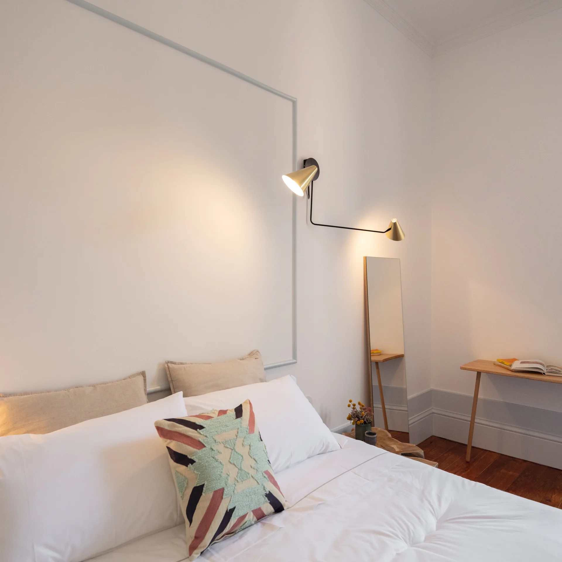 A minimalist bedroom features white bedding with a patterned pillow, a dual-arm brass wall sconce, and thin rectangular trim on the wall.