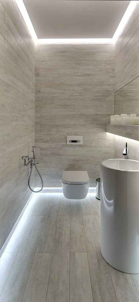 A minimalist powder room features light gray plank walls, a pedestal sink, a wall-hung toilet, and integrated floor-level LED lighting.