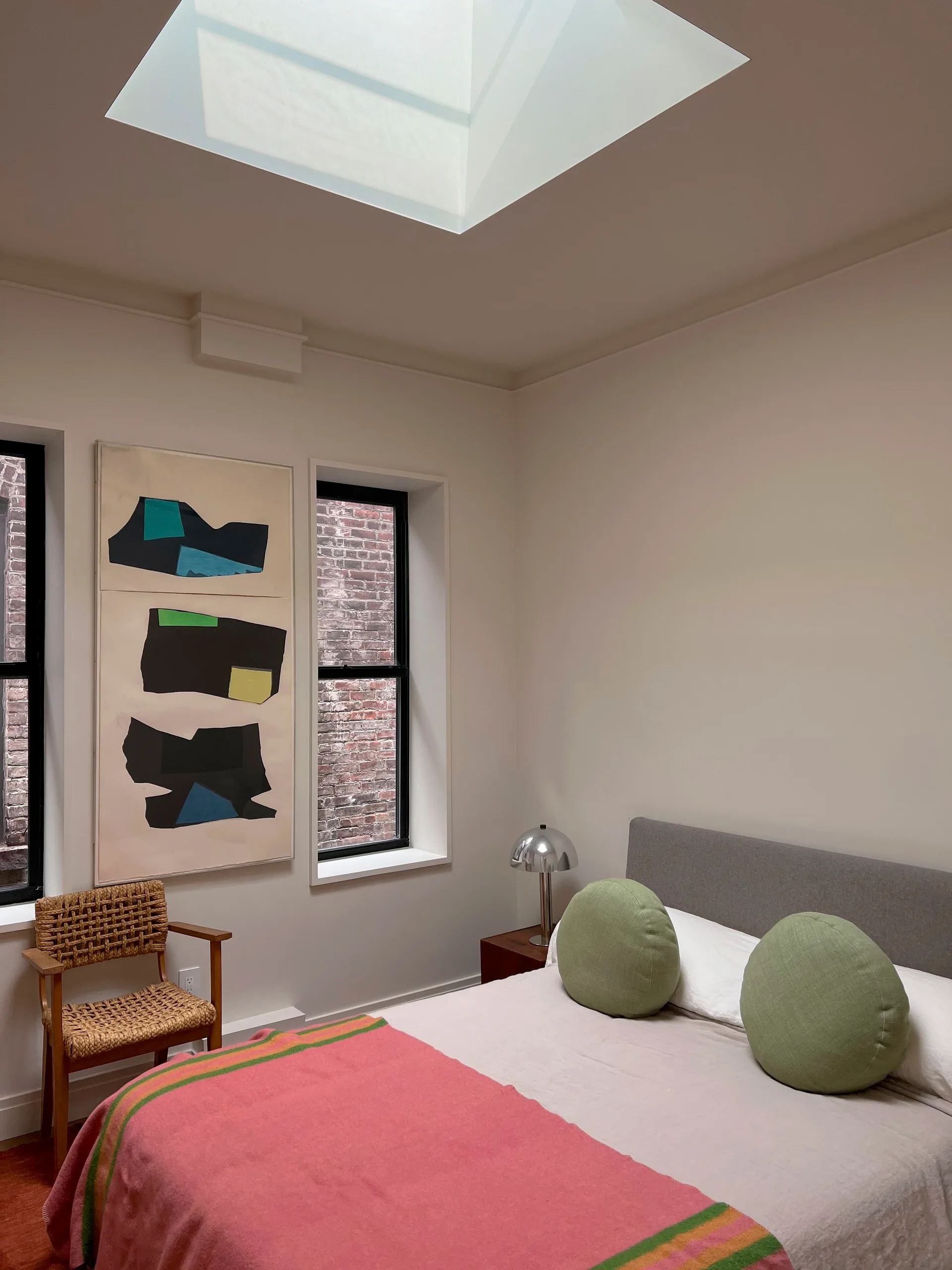 A modern bedroom features a low bed with round green pillows, a pink throw blanket, a woven accent chair, and a square skylight in the ceiling.
