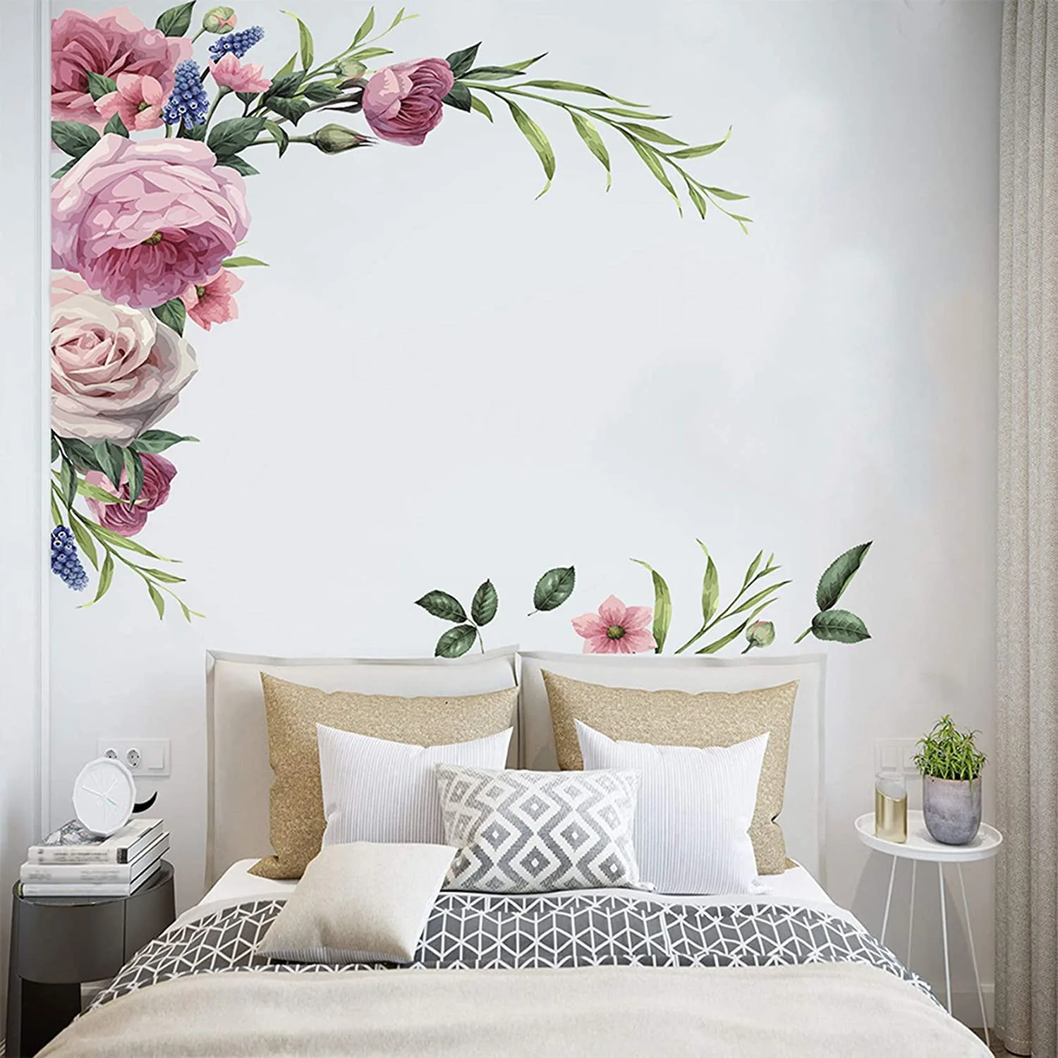 A bed with grey and beige pillows is positioned beneath a large pink floral wall decal on a plain white wall.