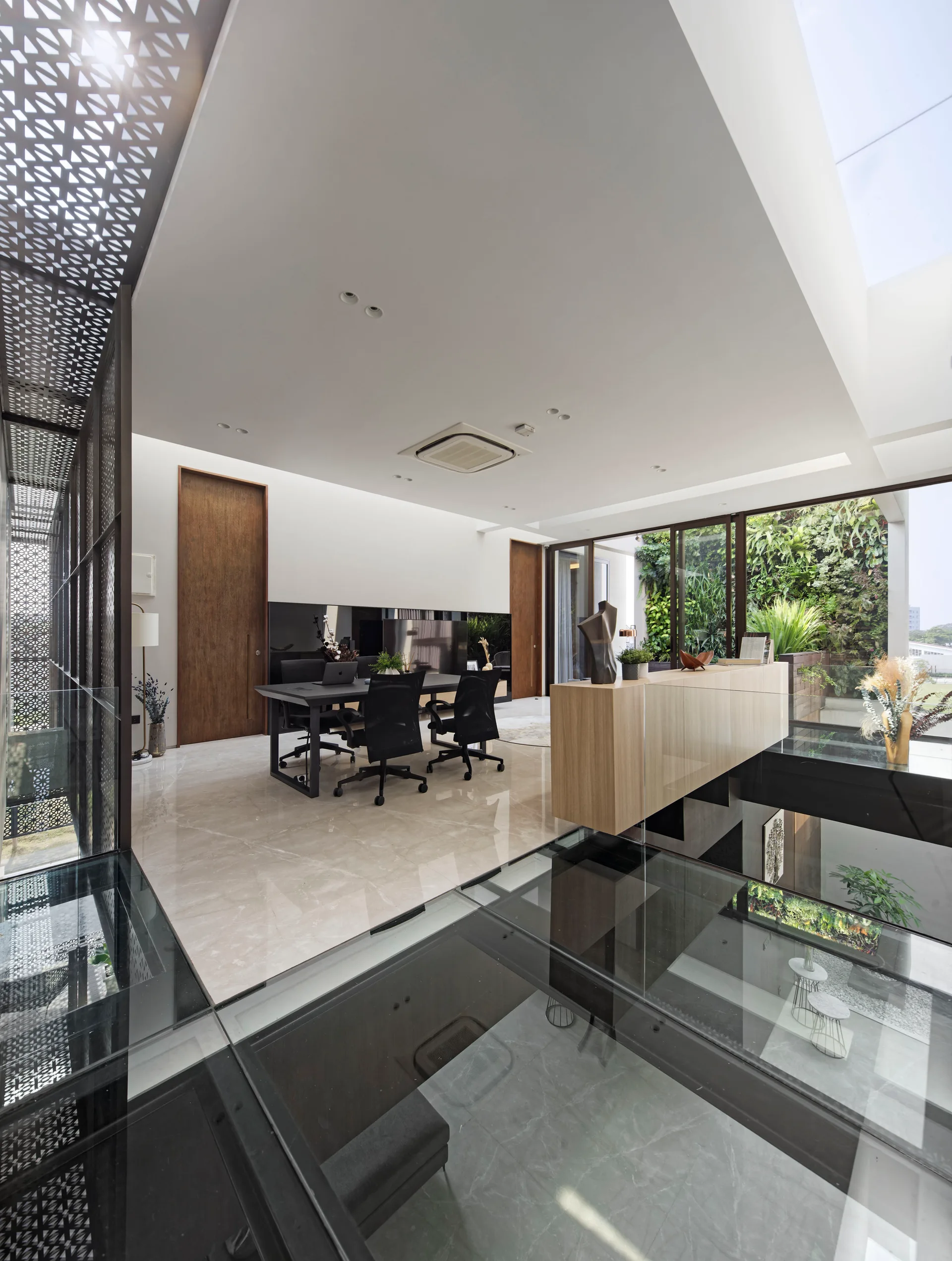 A modern home office features a black desk with office chairs on a polished tile floor next to a structural glass floor panel and a metal screen.