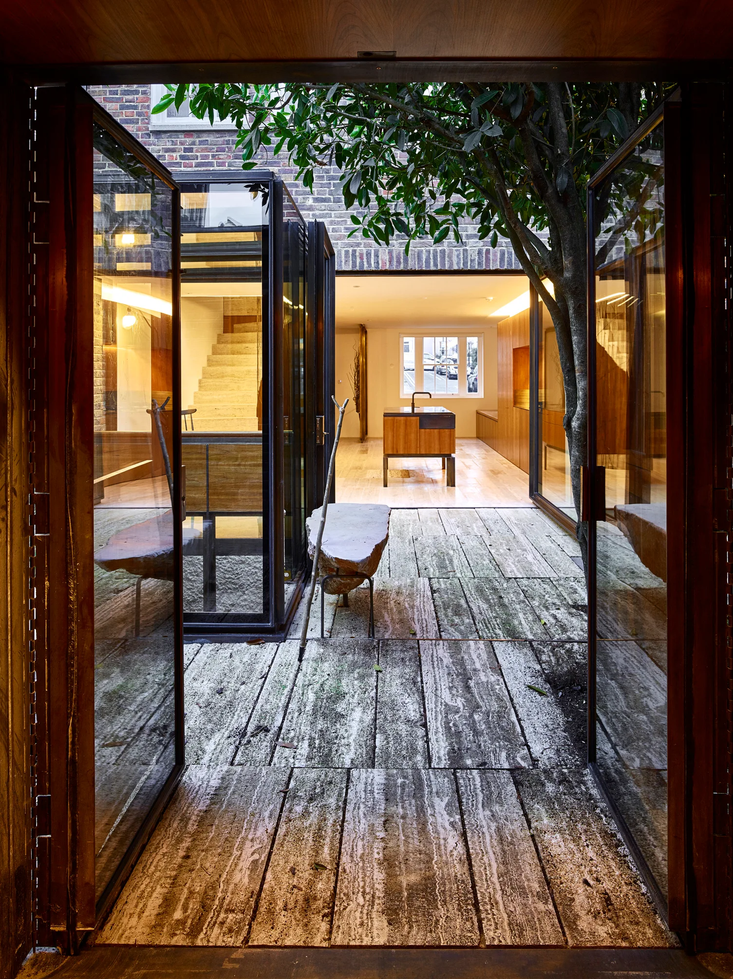 A stone-paved inner courtyard with a living tree is framed by large glass pivot doors leading to a wood-cabinet kitchen.