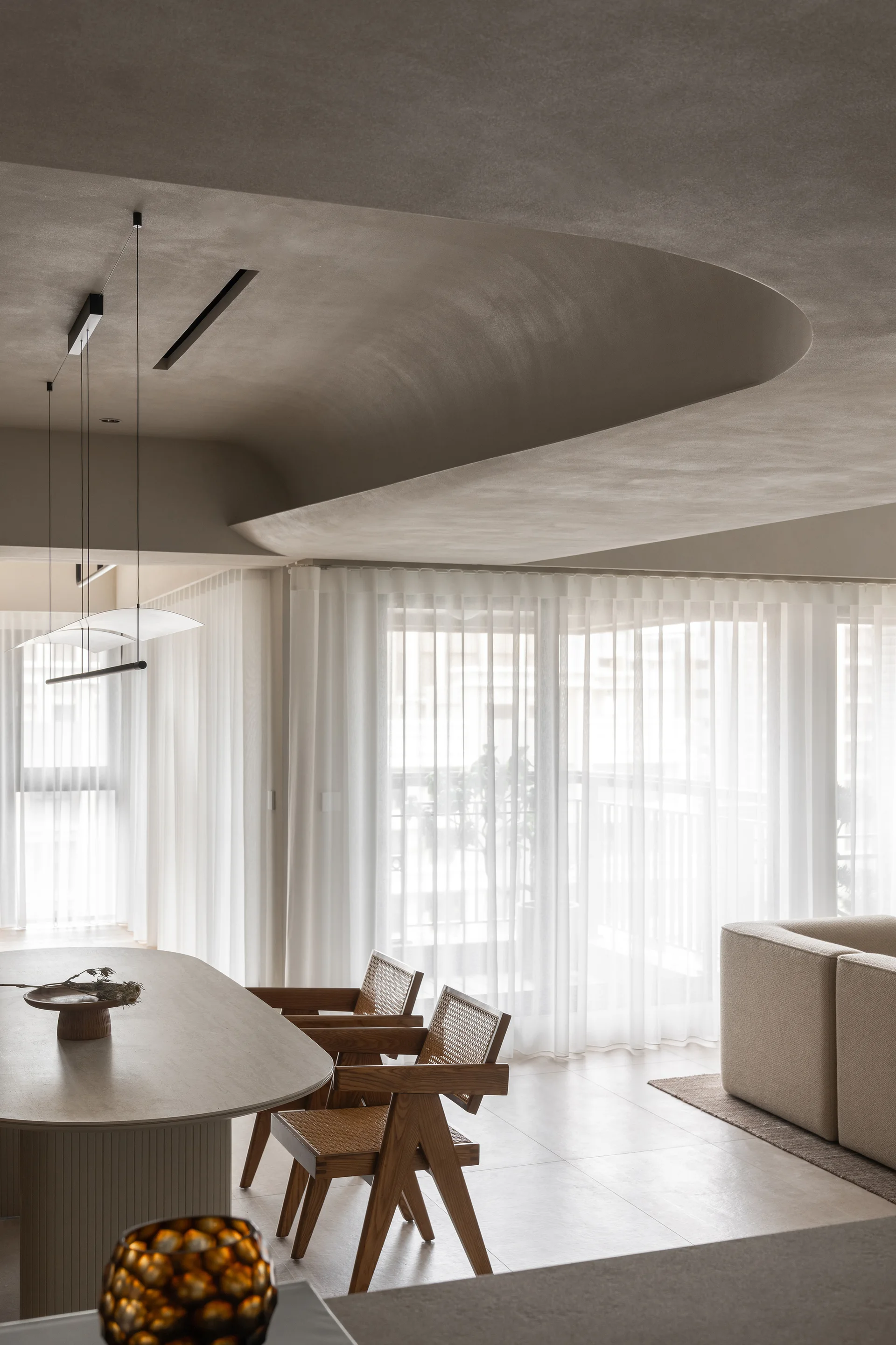 A minimalist dining space featuring a curved plaster ceiling, wooden cane-back chairs, and an oval table set against sheer curtains.