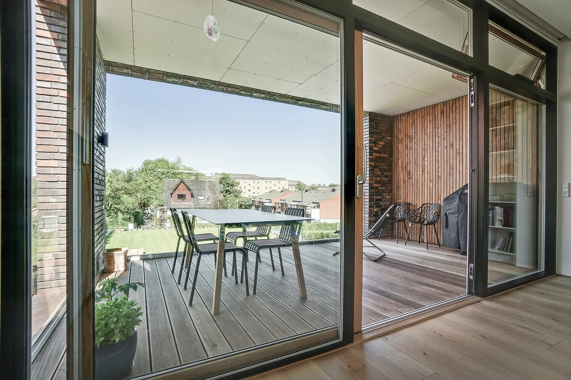 A covered balcony with wooden decking features an outdoor dining set, an exposed brick wall, and a wood-paneled accent wall behind sliding glass doors.