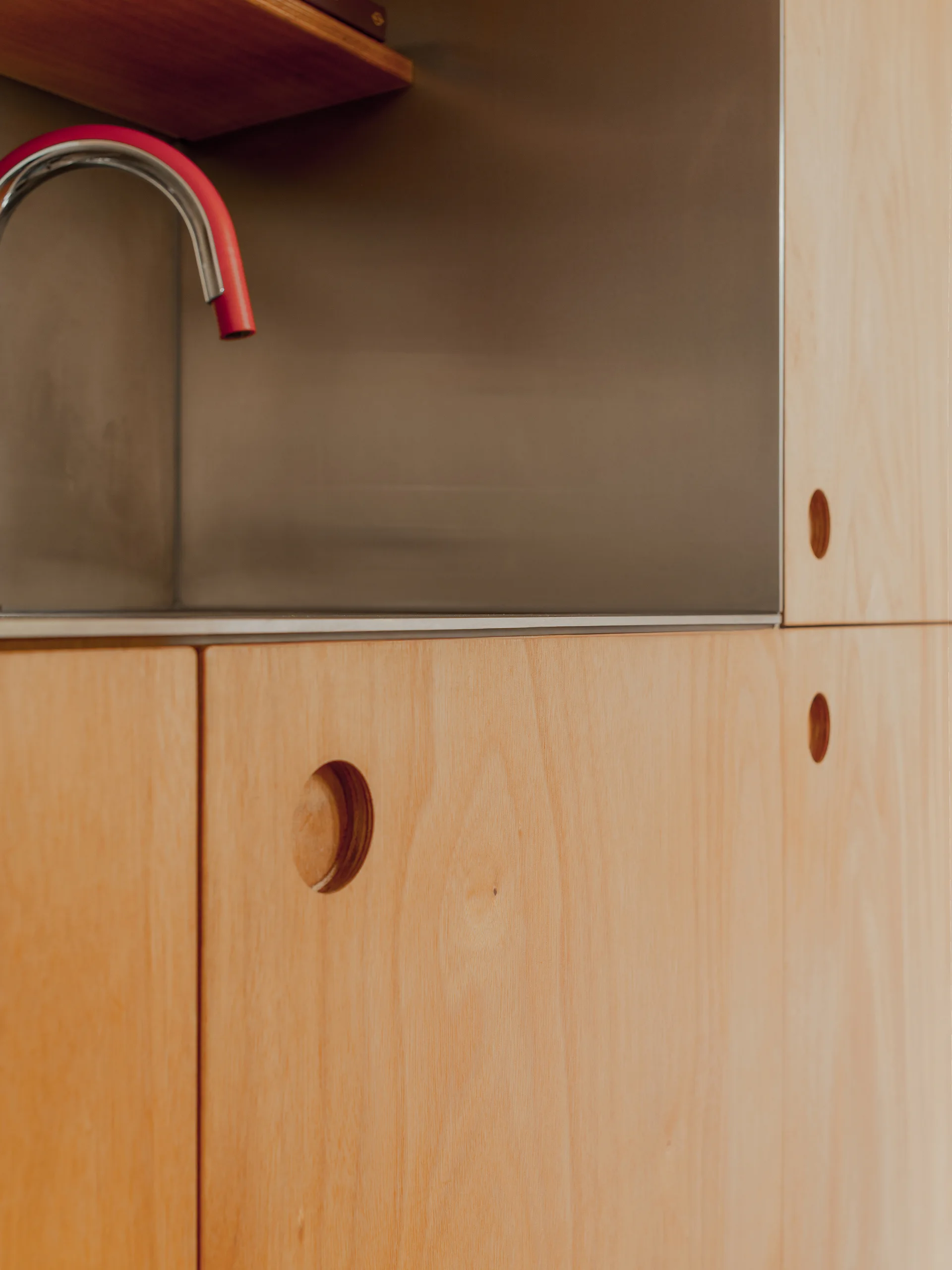 Close-up of light-toned wood kitchen cabinets with circular recessed pulls, a metal backsplash, and a curved faucet with a pink accent.