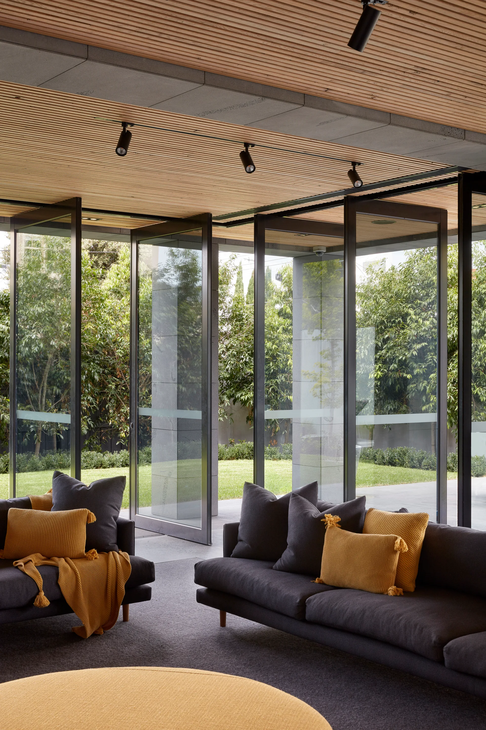 Two dark gray sofas with mustard yellow pillows sit in a living room featuring a slatted wood ceiling and pivot glass doors opening to a garden.