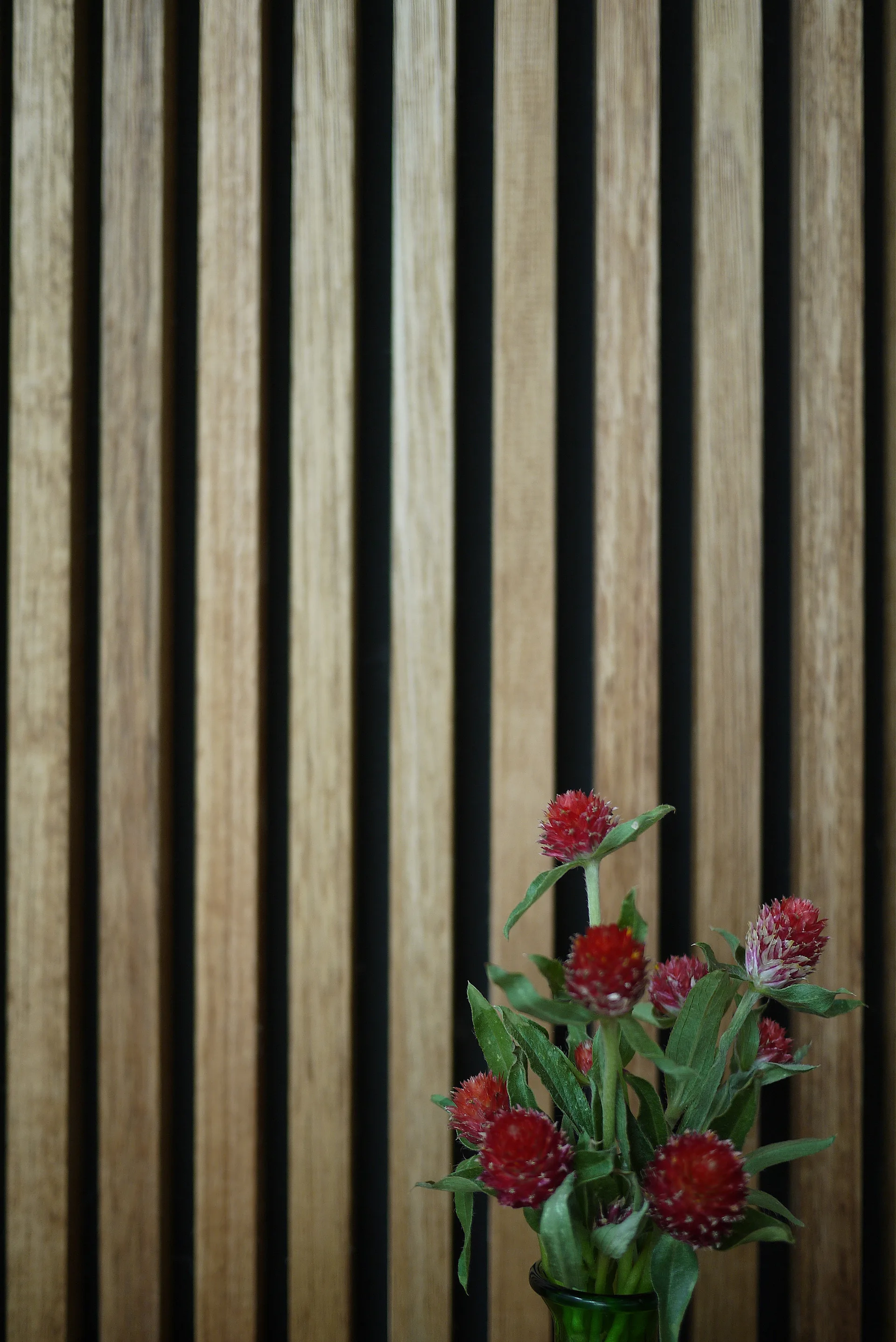 A close-up of vertical light wood slat paneling featuring a small green vase filled with round red flowers in the foreground.