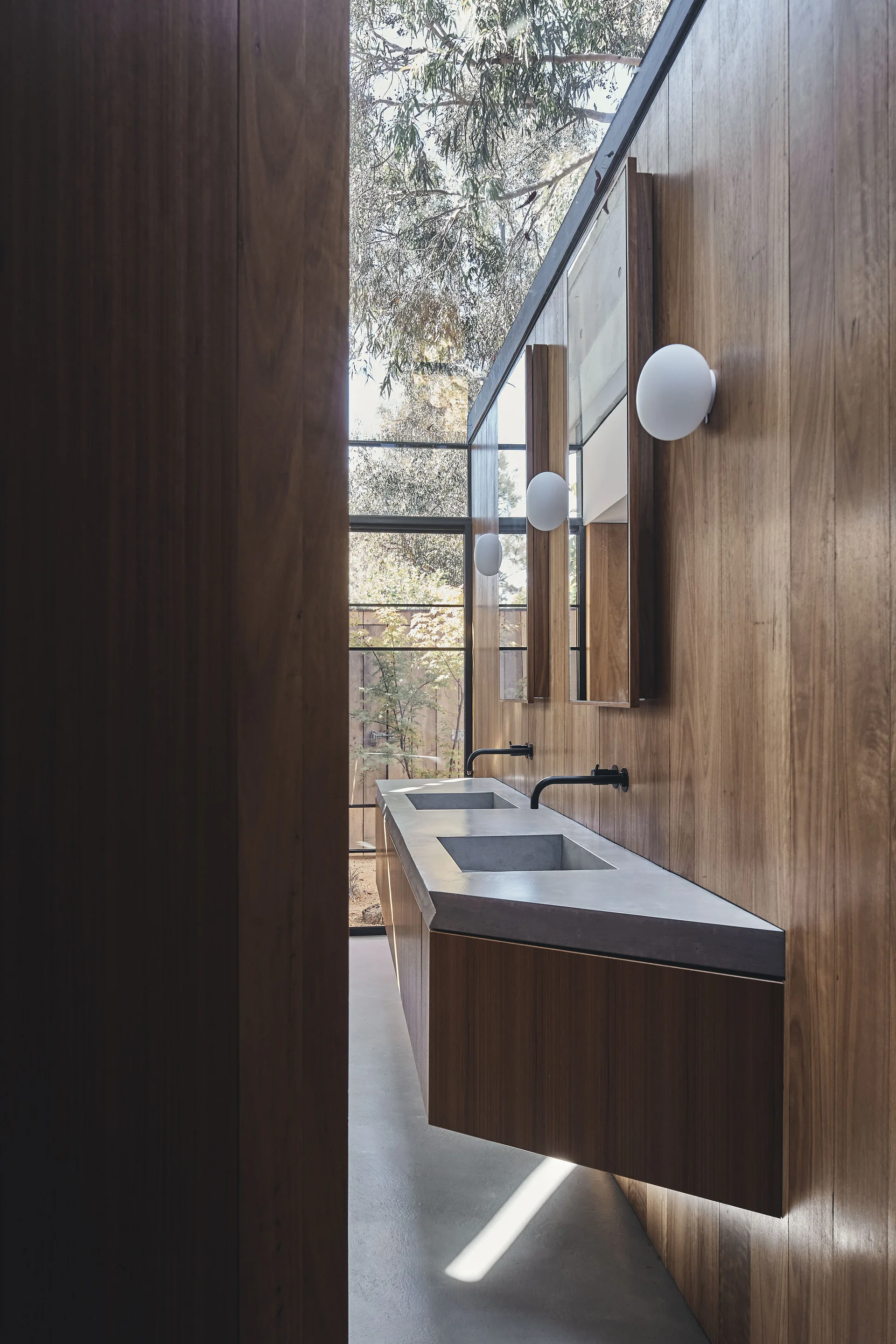 A modern bathroom with a floating wood vanity, integrated double concrete sinks, black wall-mounted faucets, and vertical wood paneling.