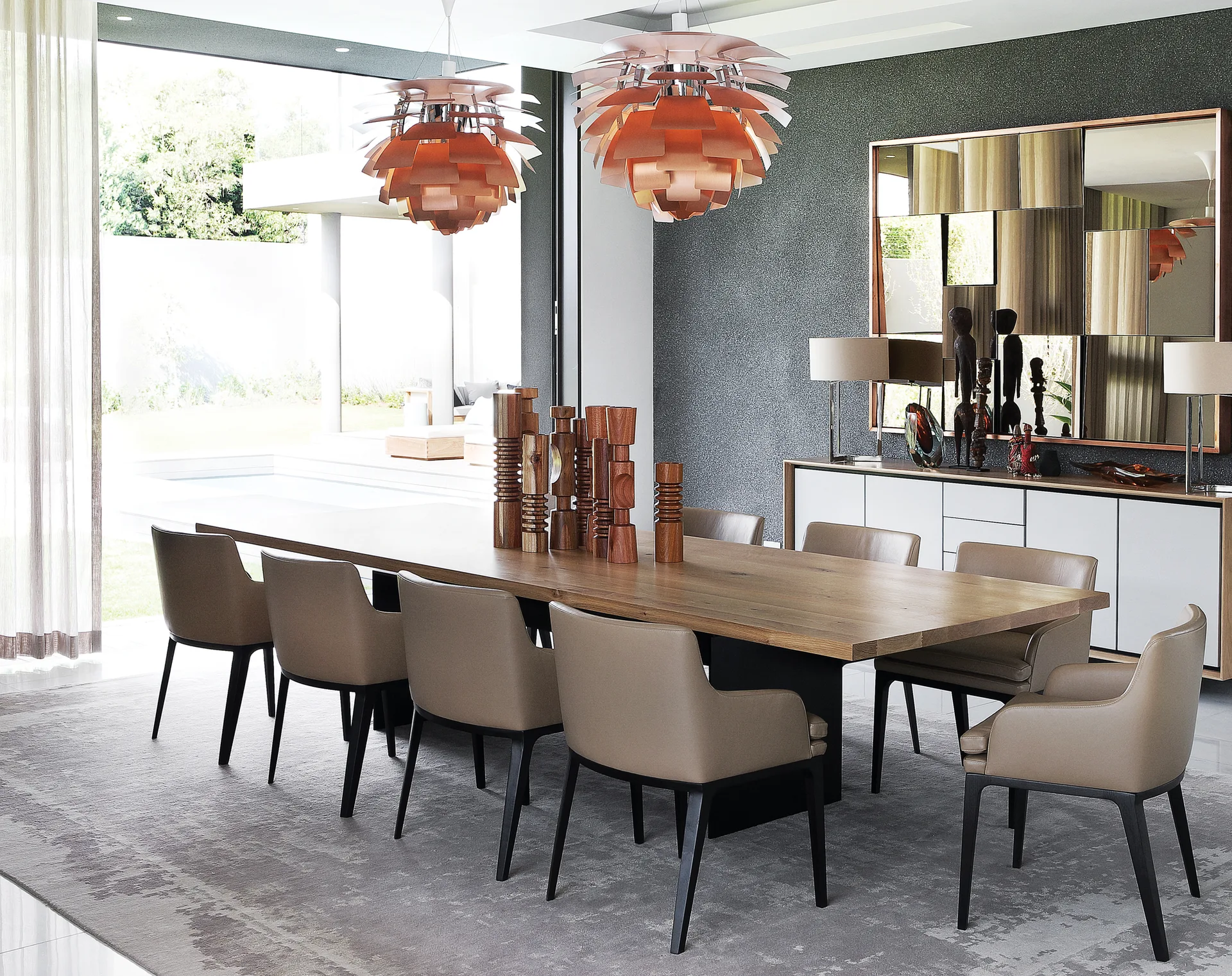 A contemporary dining room features a long wooden table surrounded by taupe chairs beneath two layered copper pendant lights.