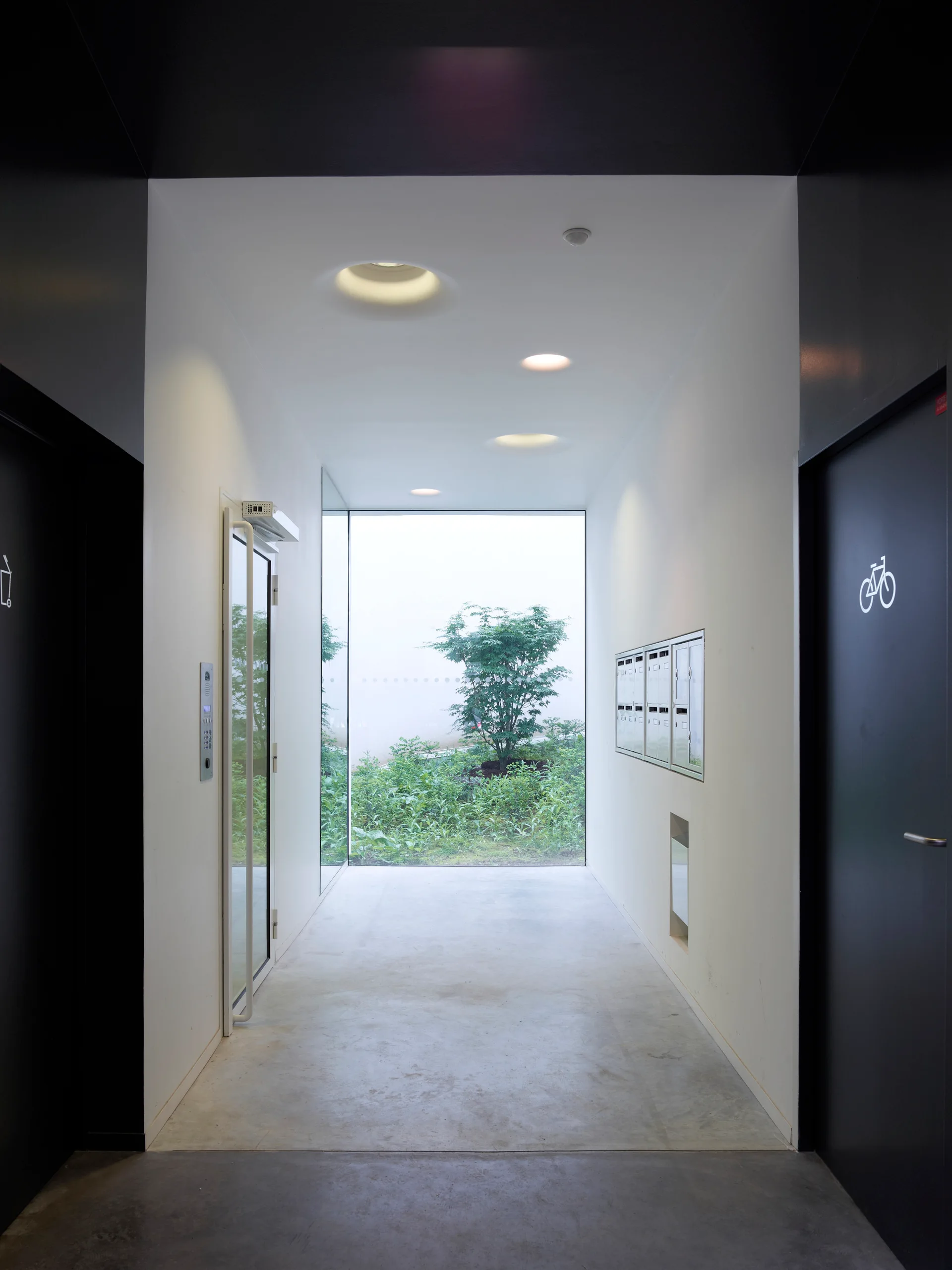 A minimalist building entryway features white walls, a concrete floor, and a large window at the end looking out at green plants.