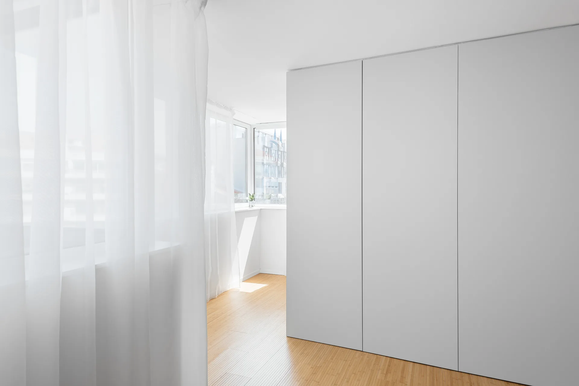 A minimalist room features a large matte white wardrobe next to light wood flooring and windows covered with sheer white curtains.