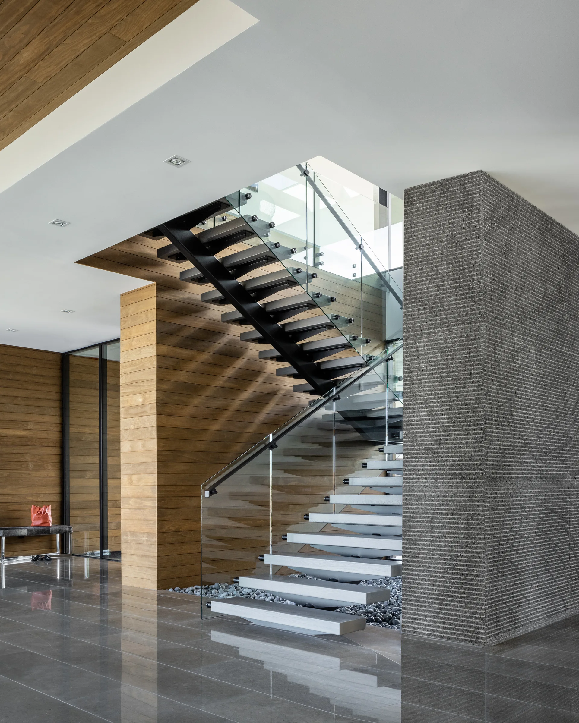 A modern staircase with open risers and glass railings is flanked by a warm wood slatted wall and a textured gray stone column.