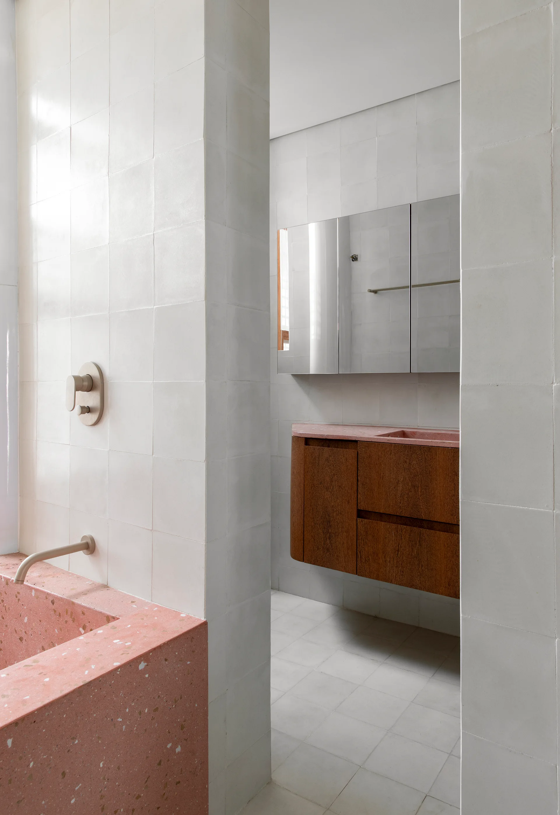 A modern bathroom features a pink terrazzo bathtub and vanity countertop with a floating wood cabinet against white tiled walls.