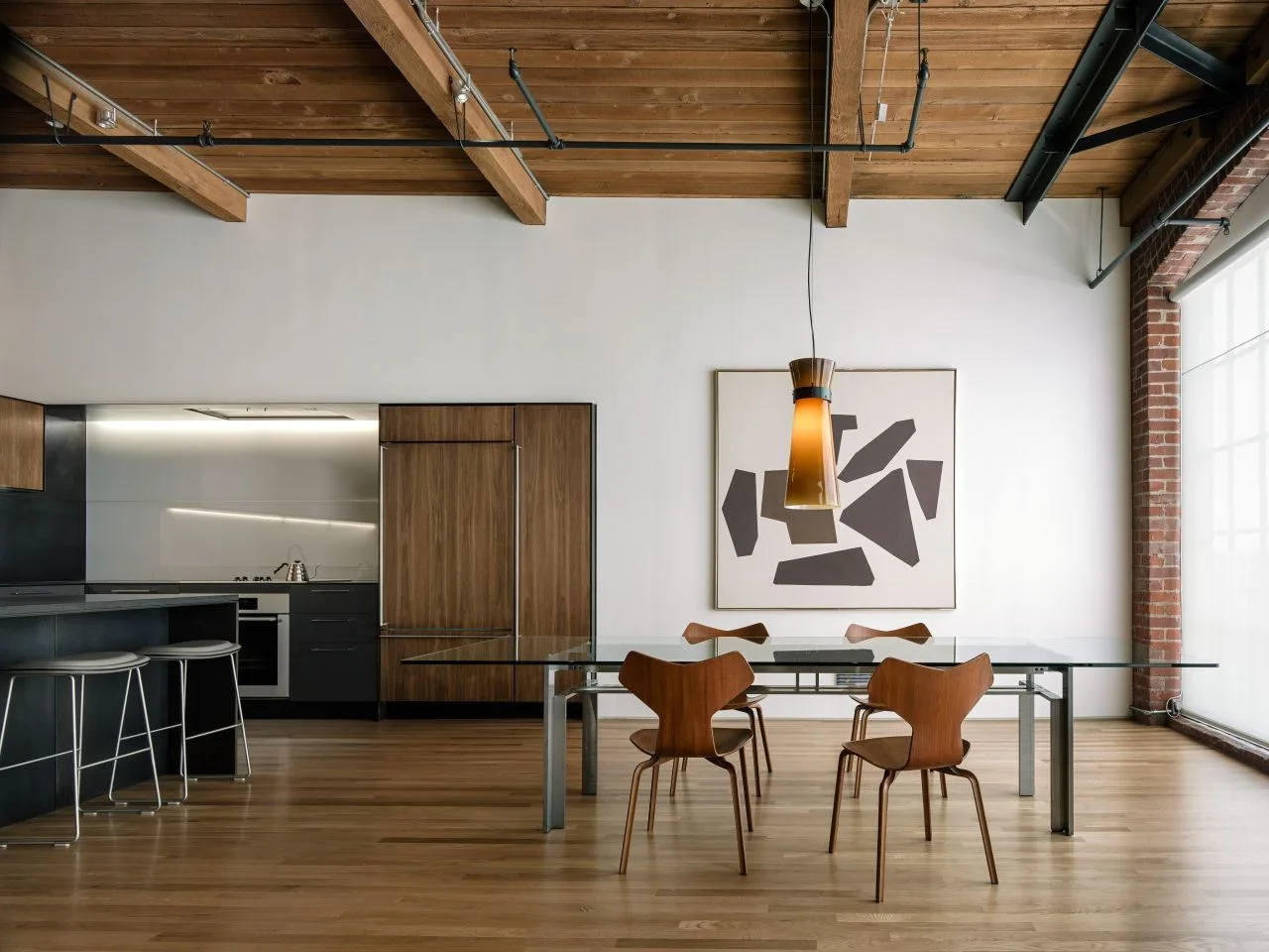 A modern loft dining area featuring a glass-top table, molded wooden chairs, an amber pendant light, and exposed wooden ceiling beams.