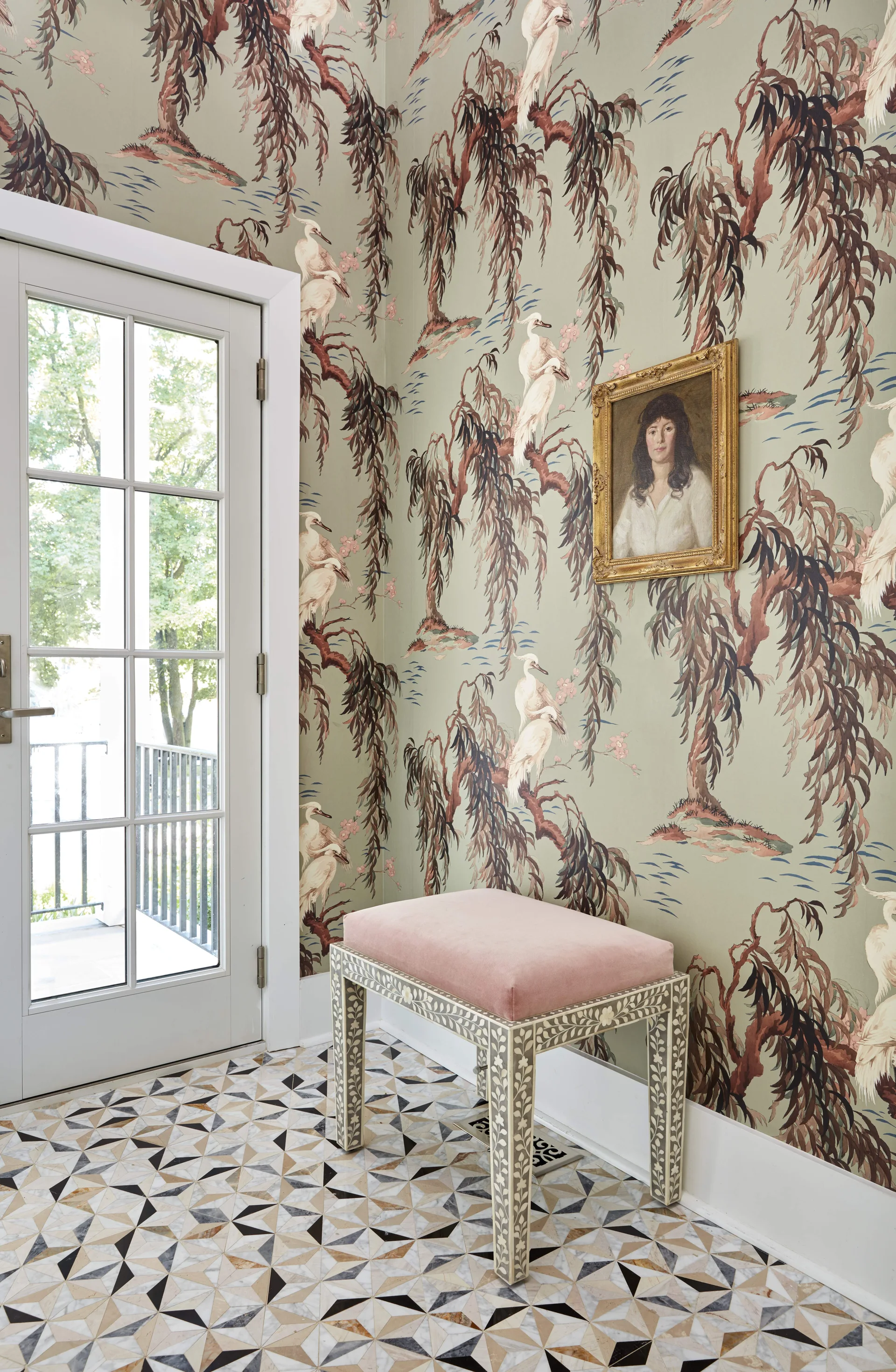 An entryway featuring green botanical wallpaper, a pink upholstered bench with patterned legs, and a geometric tiled floor next to a white door.