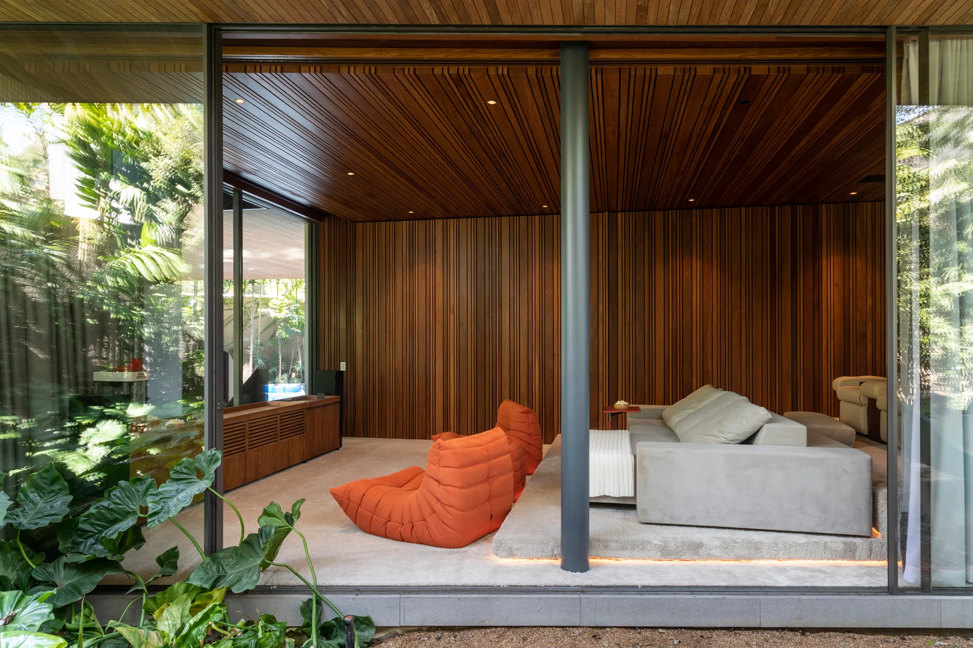 A modern lounge room features vertical wood slat walls and ceiling, a gray sectional sofa on a carpeted platform, and two orange lounge chairs.