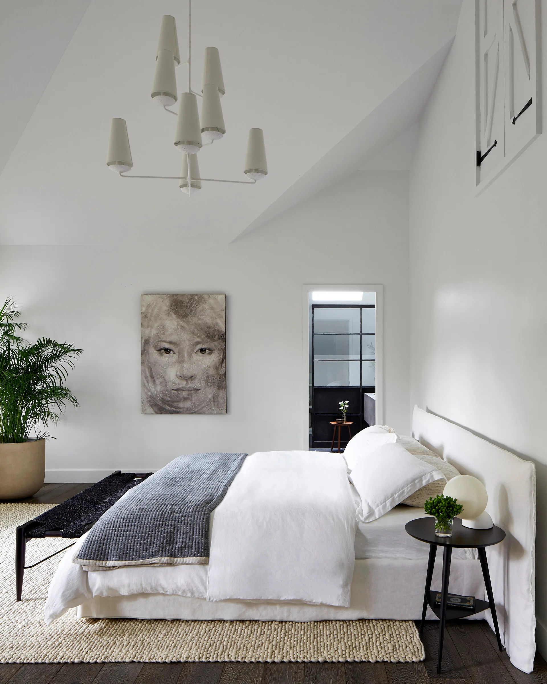 A minimalist white bedroom with a vaulted ceiling, a textured woven rug, a white bed, and a large black-and-white portrait painting.