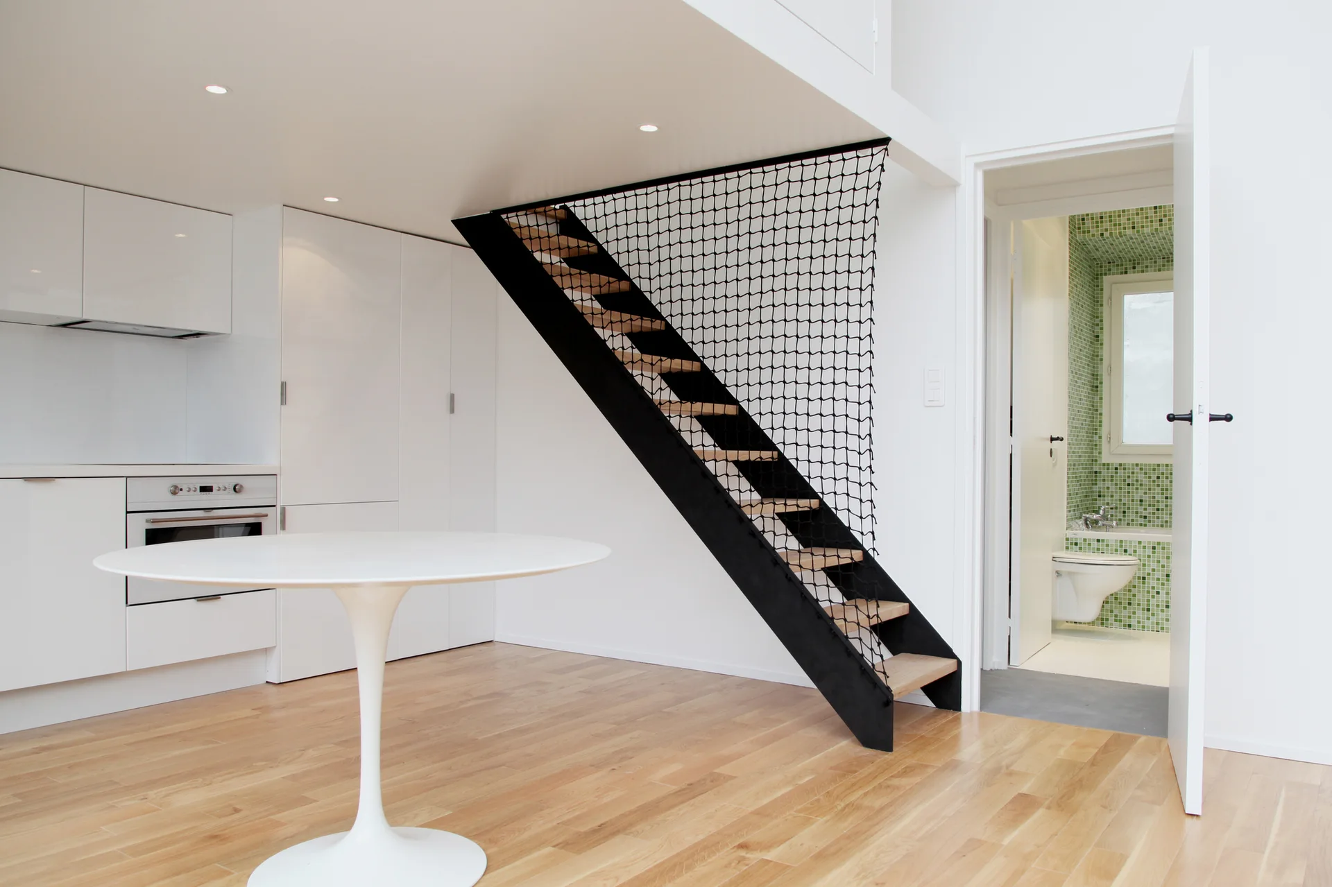 A minimalist kitchen with a white pedestal table next to a black staircase with safety netting, with a glimpse of a green-tiled bathroom.