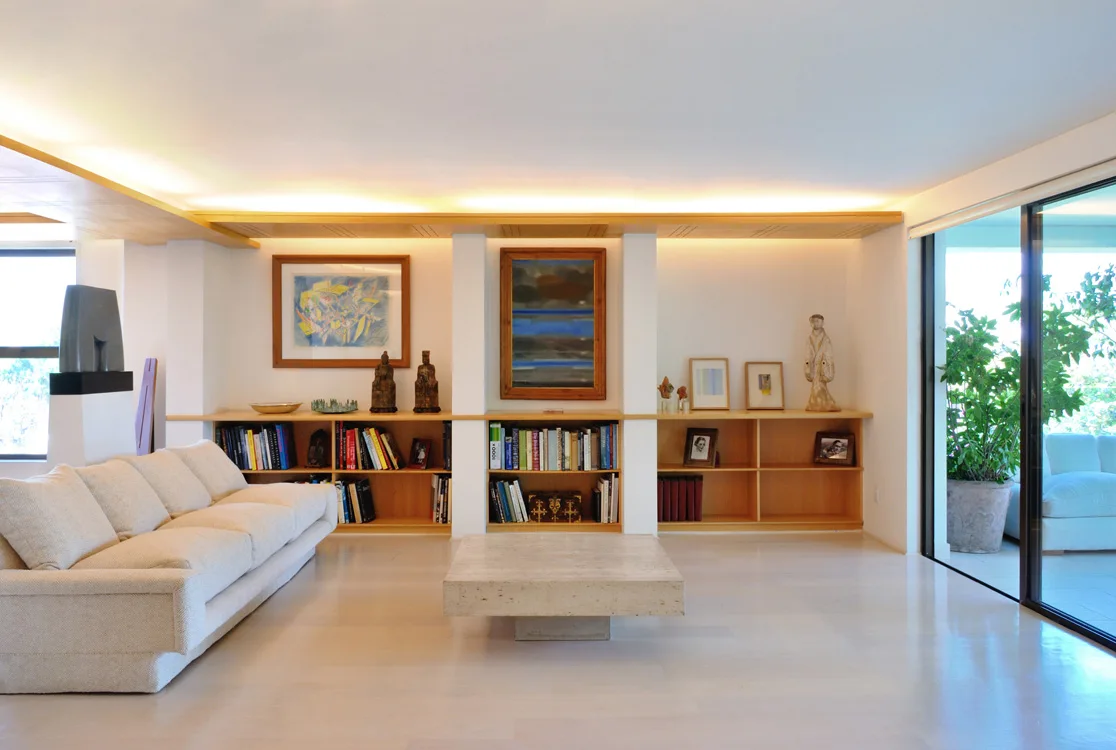A spacious living room features a cream sofa, a square stone coffee table, and low wood built-in bookshelves illuminated by integrated cove lighting.