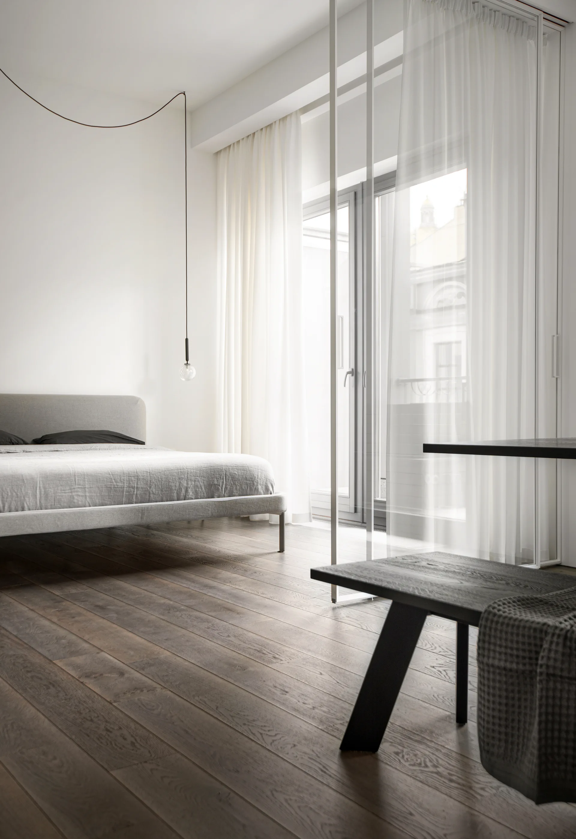 A minimalist bedroom featuring a grey bed on dark hardwood floors, next to a white-framed glass partition and sheer white curtains.