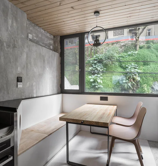 A minimalist dining nook features a wooden table, built-in bench, concrete wall, and a large window overlooking green foliage.
