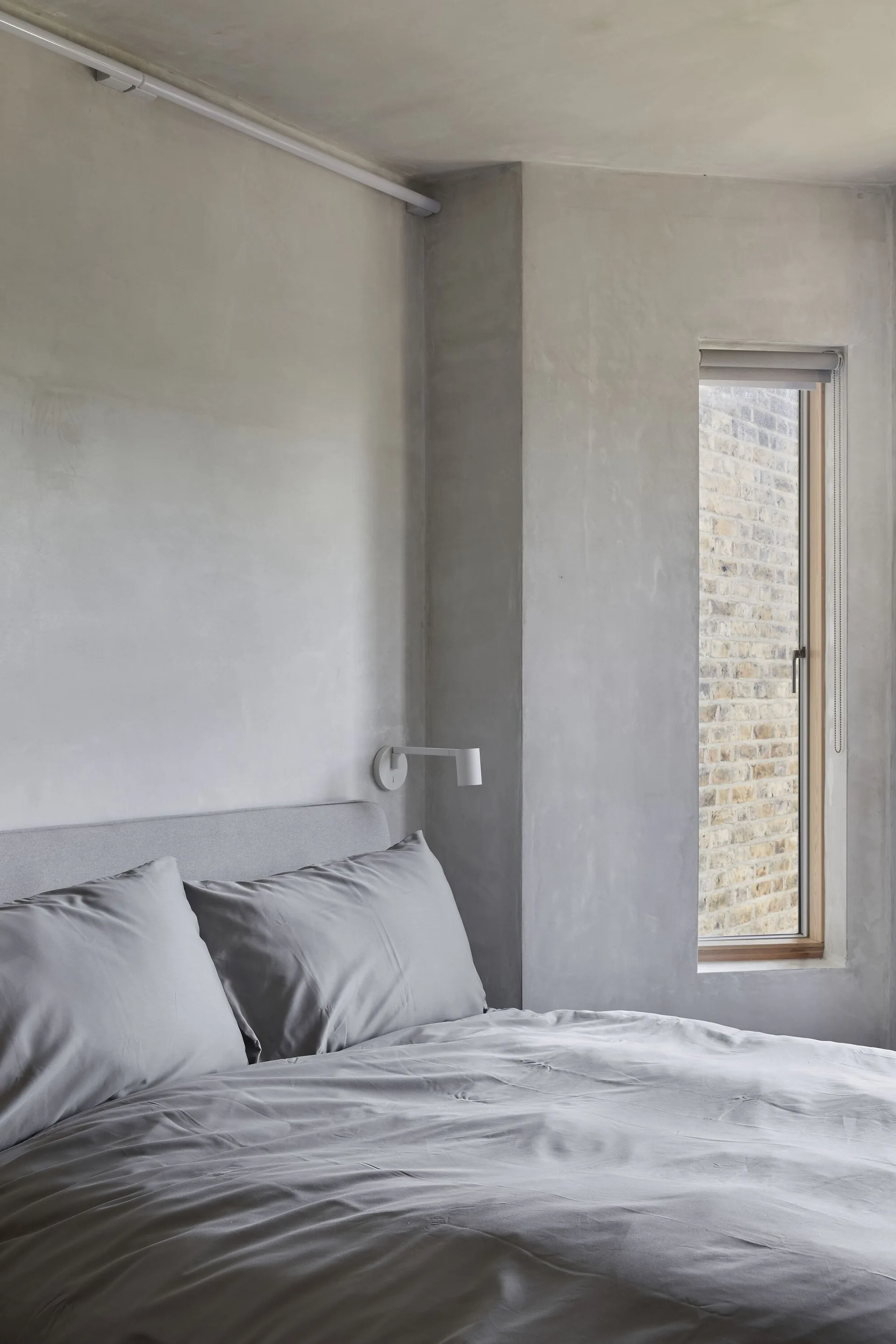 A minimalist bedroom features textured grey plaster walls, a bed with grey sheets, a white wall-mounted reading lamp, and a narrow recessed window.