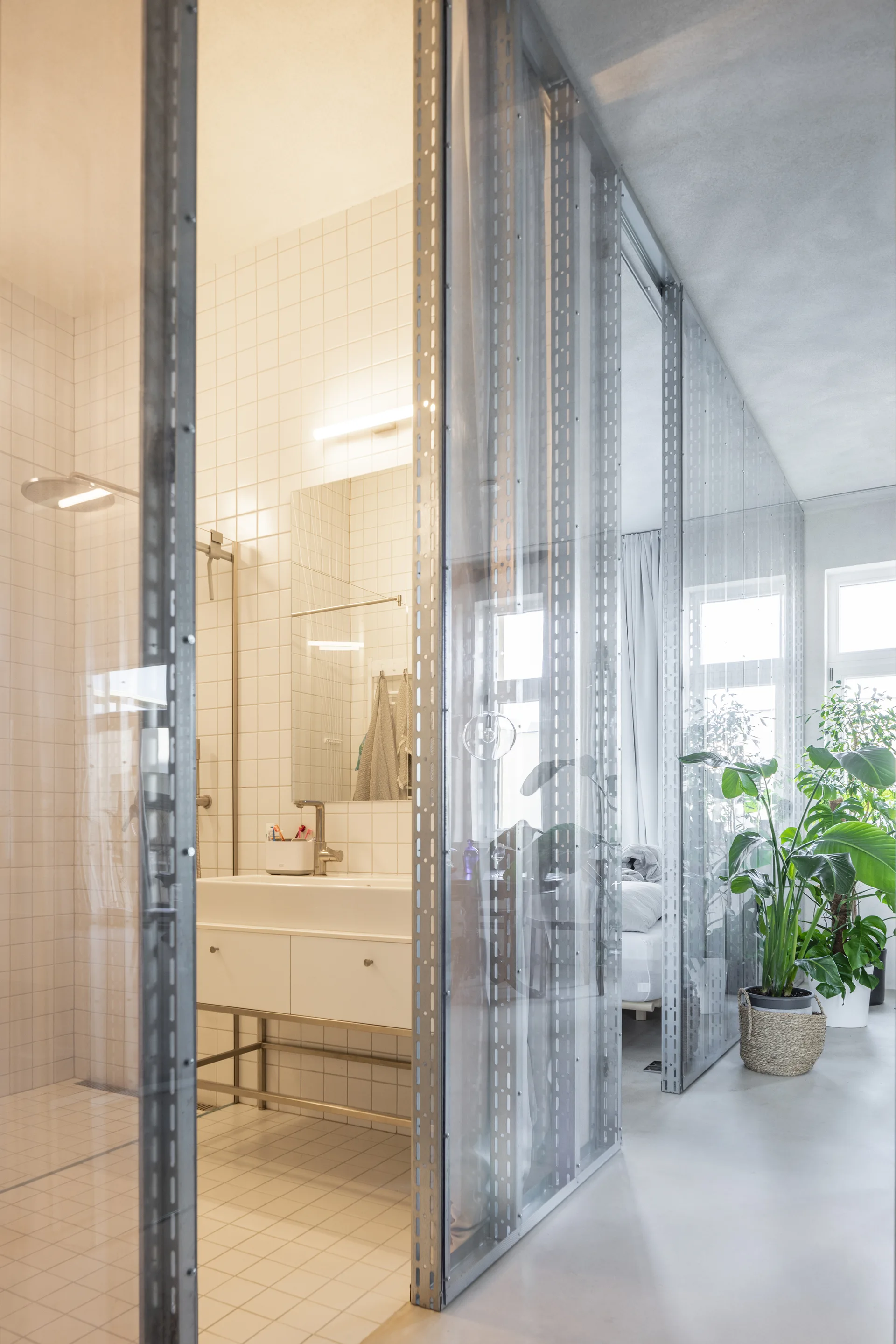 A white tiled bathroom with a minimalist vanity is separated from a living space by a metal-framed glass partition wall.