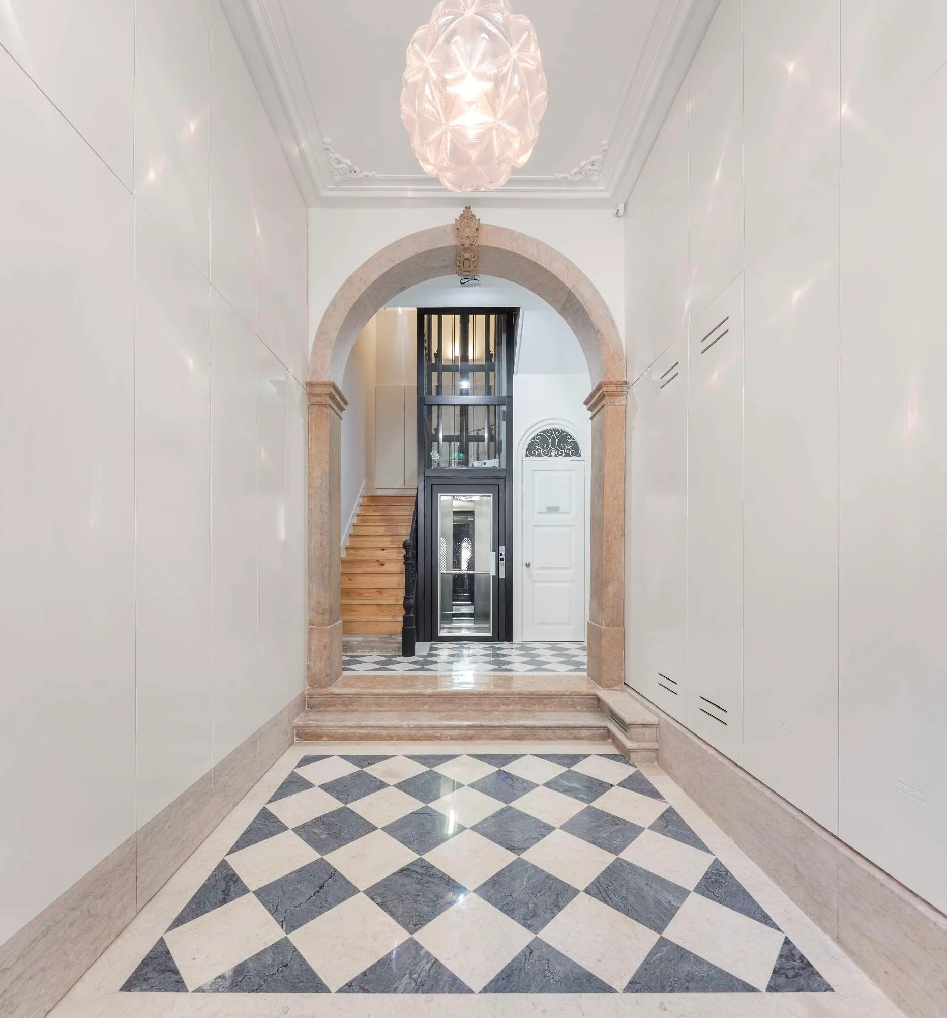 A foyer featuring a checkerboard marble floor, glossy white walls, and a central stone archway leading to a modern glass elevator.