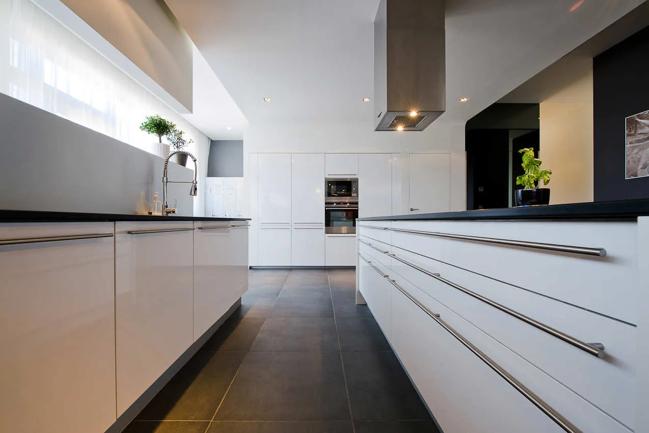 A modern kitchen features glossy white slab cabinets with long metal bar pulls, dark countertops, and a large format grey tile floor.