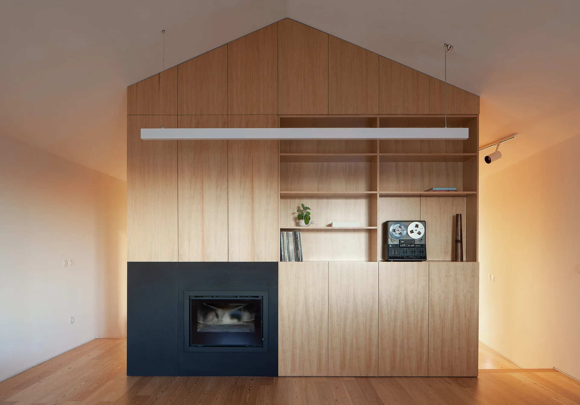 A floor-to-ceiling light wood storage unit built into a gabled wall, featuring open shelving, a black fireplace, and a hanging linear light.
