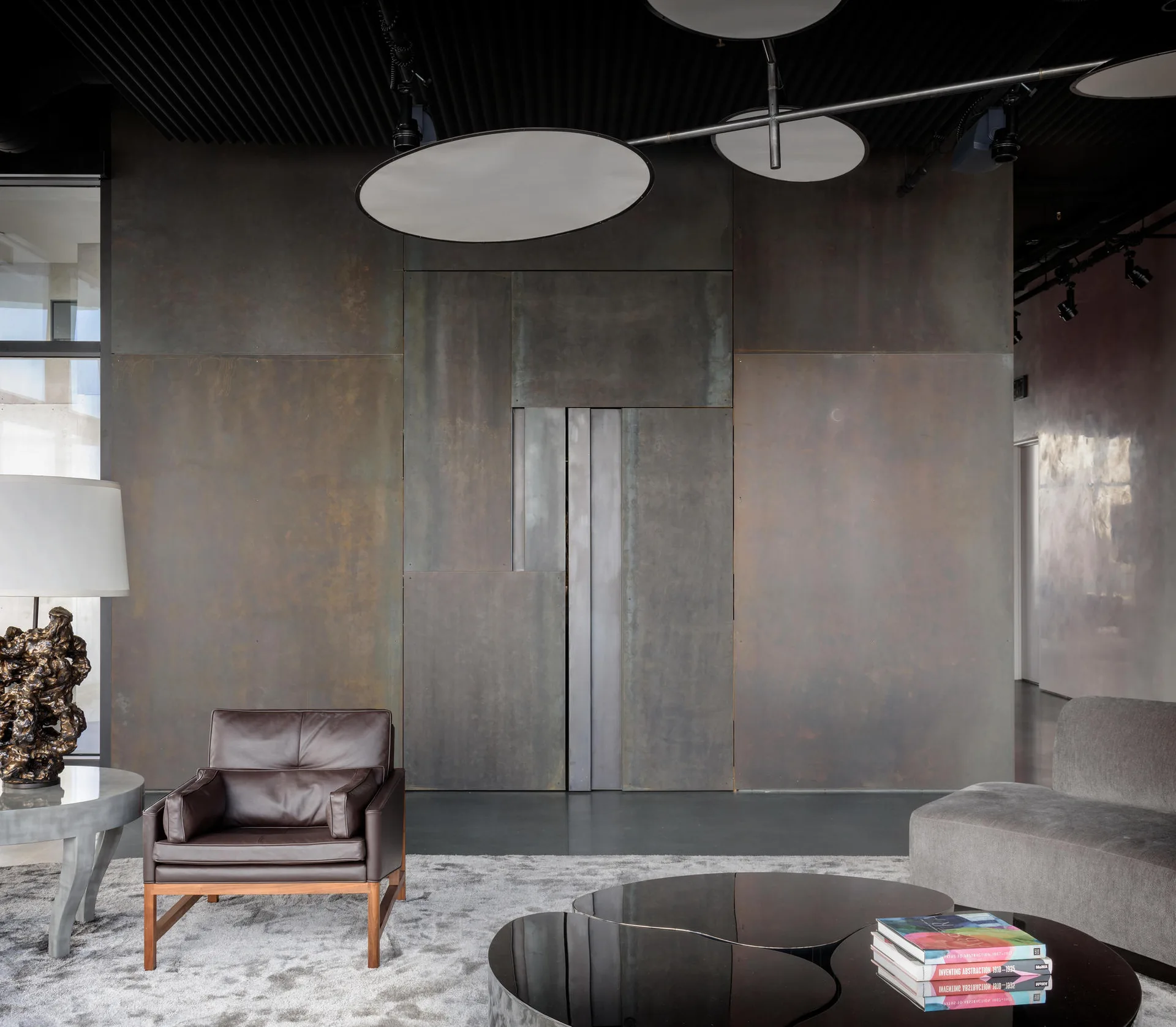 An industrial living room featuring oxidized metal wall panels, a dark brown leather armchair, and a black organic-shaped coffee table.