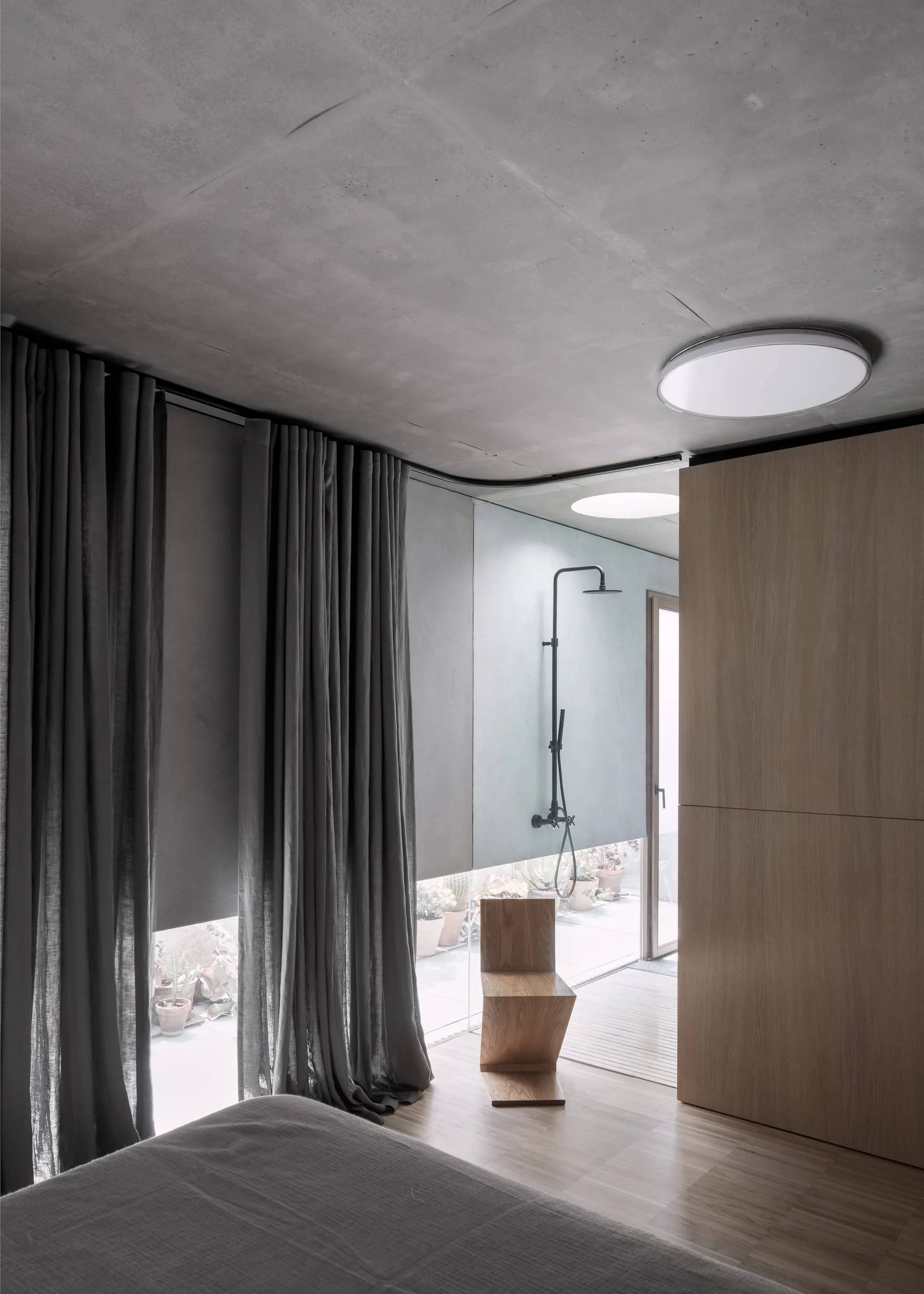 A minimalist bedroom featuring an exposed concrete ceiling, a sculptural wooden chair, grey drapes, and an open glass shower in the background.
