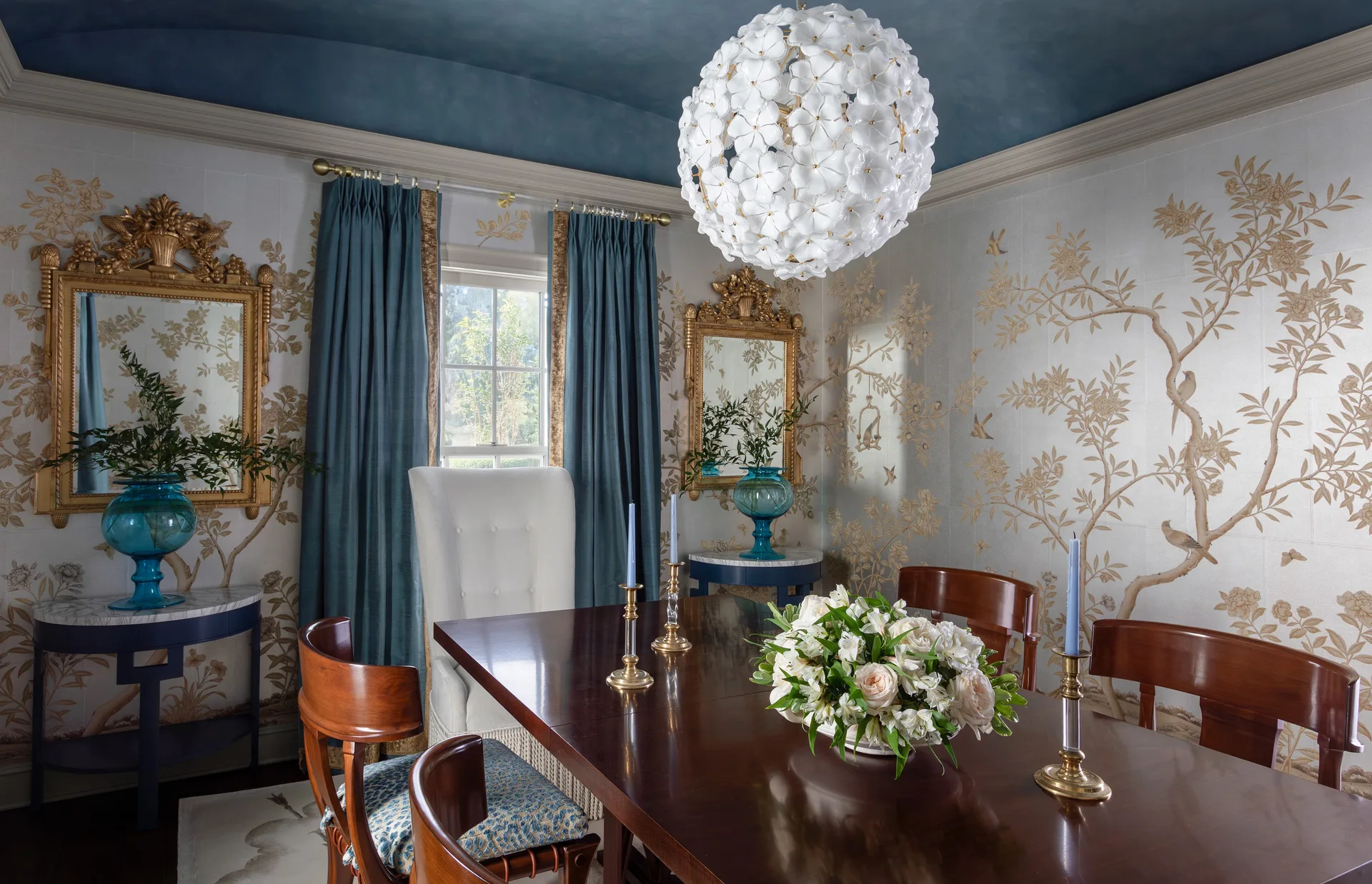 A formal dining room features a dark wood table, floral wallpaper, ornate gold mirrors, and a white spherical floral chandelier hanging from a blue ceiling.