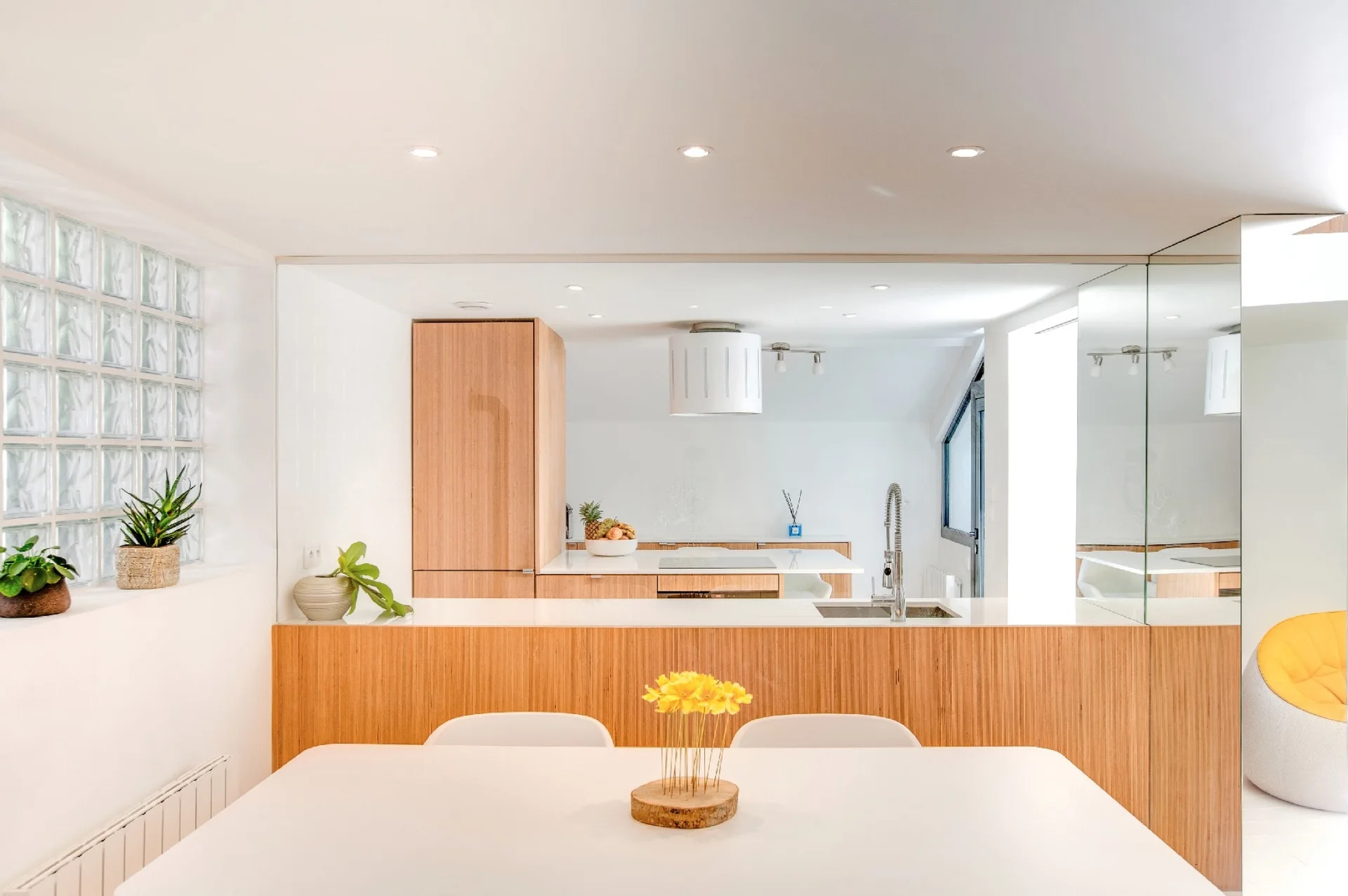 A minimalist kitchen and dining area features white countertops, wood-paneled cabinetry, a dining table with yellow flowers, and a glass block wall.