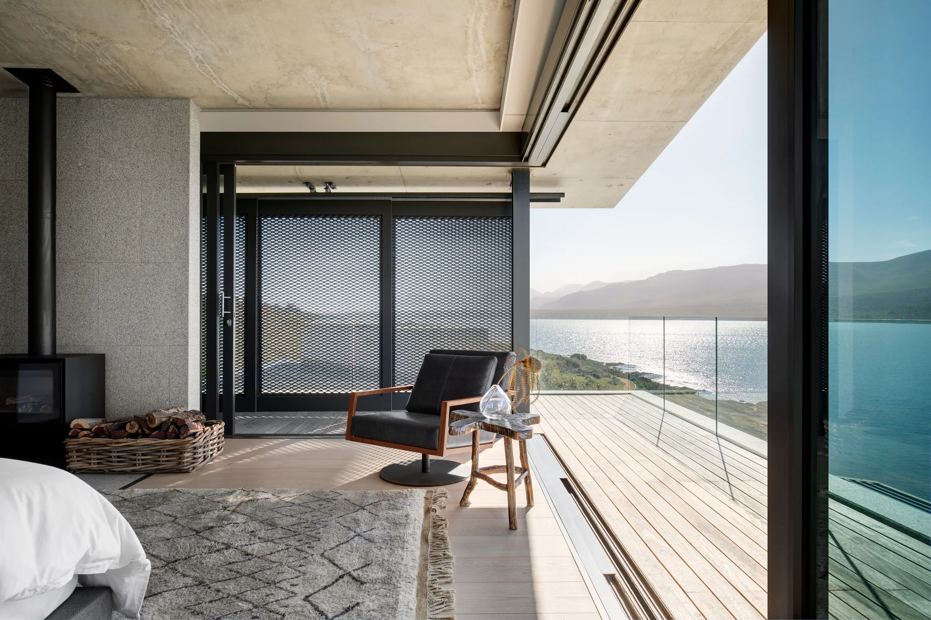 A modern bedroom features an exposed concrete ceiling, a black lounge chair, and large sliding glass doors opening to a wooden deck with a scenic lake view.