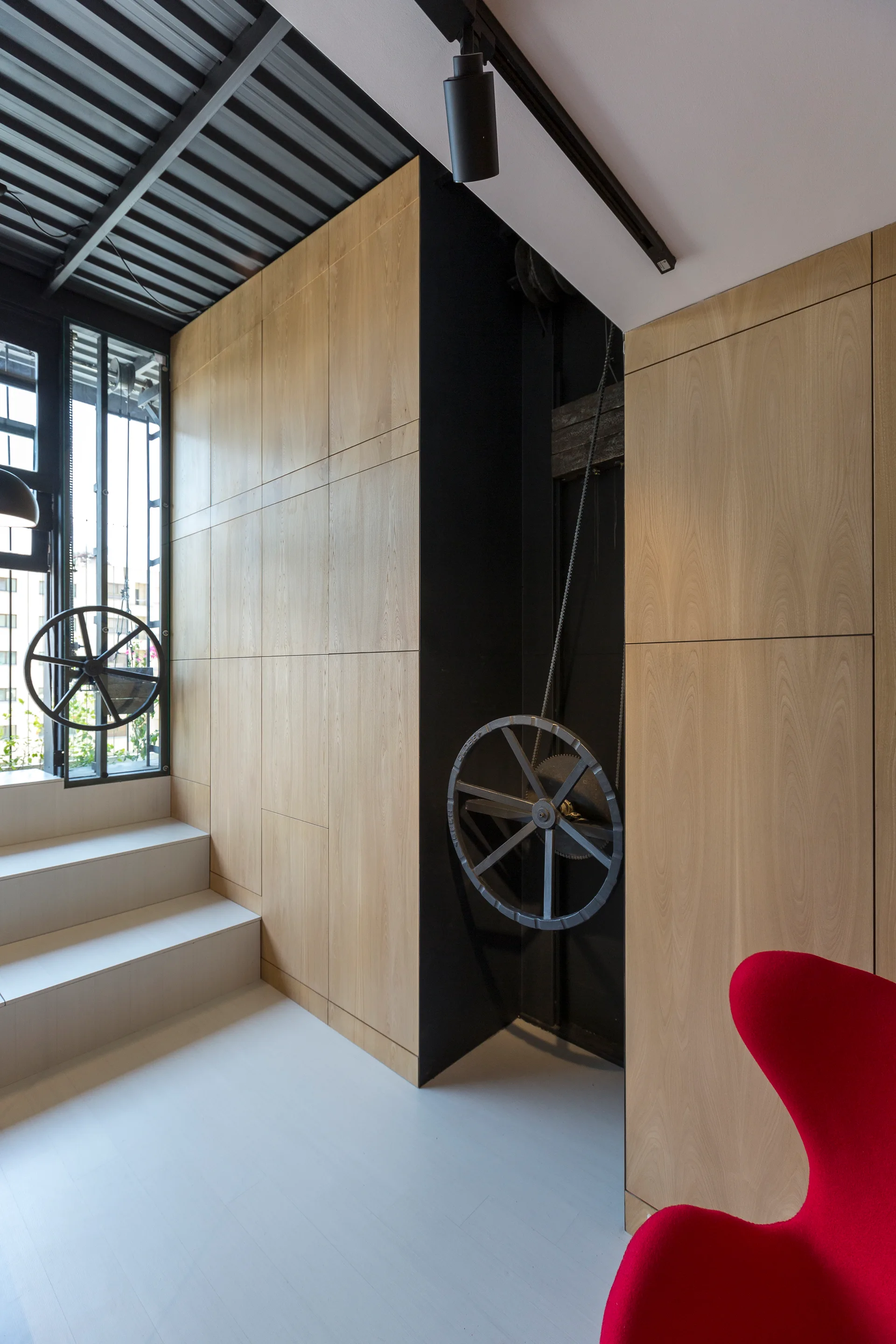 A modern room features a tall light wood storage wall, grey stepped platforms leading to a window, and decorative black gear wheels with chains.